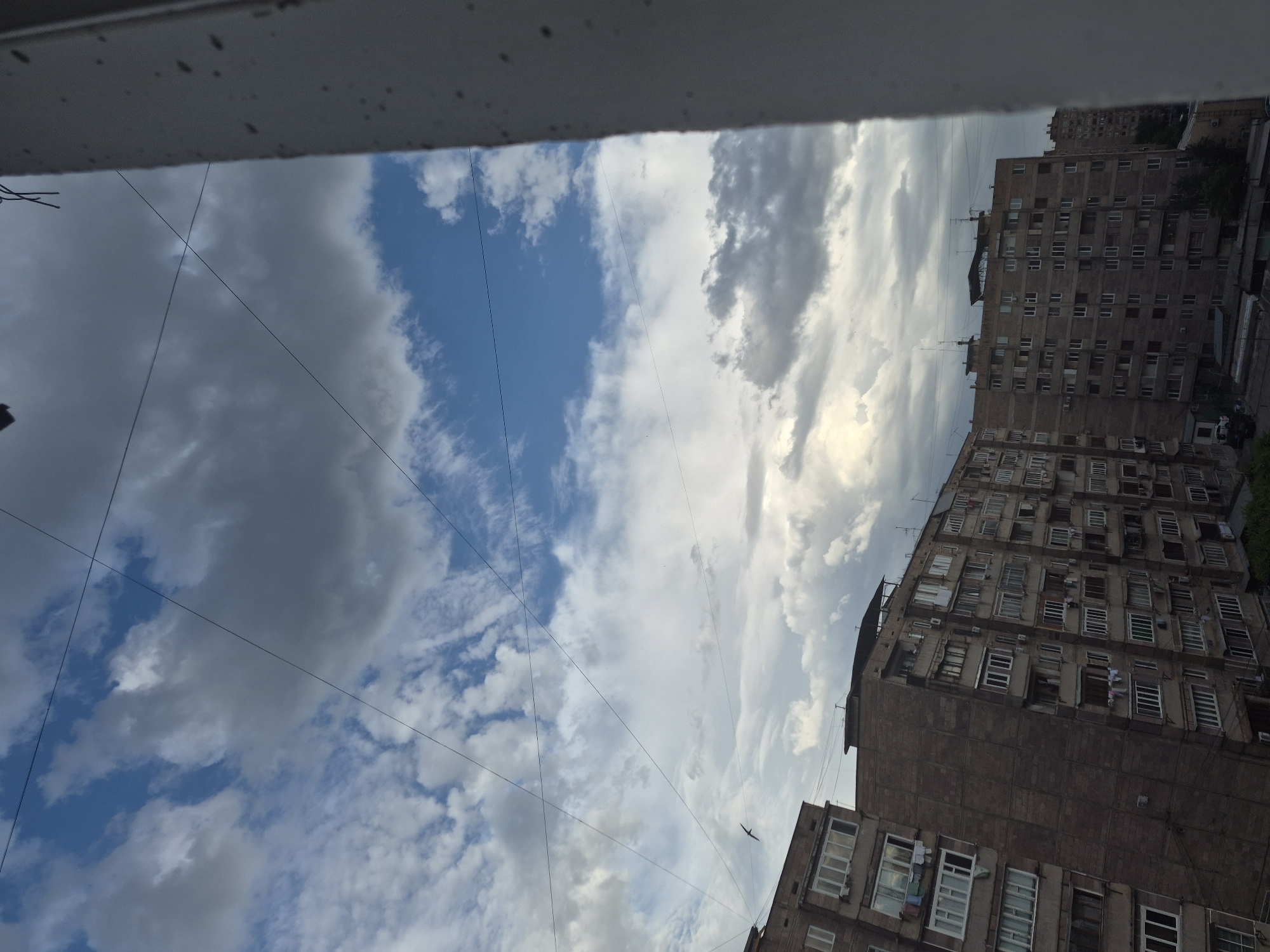 The image, viewed in its correct upright orientation, presents an urban landscape primarily featuring a dynamic sky and residential buildings. The upper portion of the frame is obscured by a dark, blurry structure, likely the edge of a window or balcony from which the photograph was taken.
The sky is a dominant element, showcasing a varied cloudscape with patches of clear blue sky interspersed with both dark grey and brightly illuminated white clouds. A strong light source, presumably the sun, is visible breaking through the clouds on the upper right side, suggesting either early morning or late afternoon.
Below the sky, two large, multi-story residential buildings made of a dark, weathered concrete or stone material are prominent. These buildings, typical of Soviet-era architecture, are densely packed with numerous windows, some open, and others displaying air conditioning units or clothes drying on balconies. Their functional design and weathered appearance are consistent with the urban environment of Yerevan, Armenia.
Several thin, dark utility wires crisscross the sky between the buildings. A small, dark bird is captured in flight against the bright sky on the right side of the image. The scene conveys a sense of everyday urban life on a partly cloudy day. No specific human activity is visibly occurring, and no text is discernible in the image.