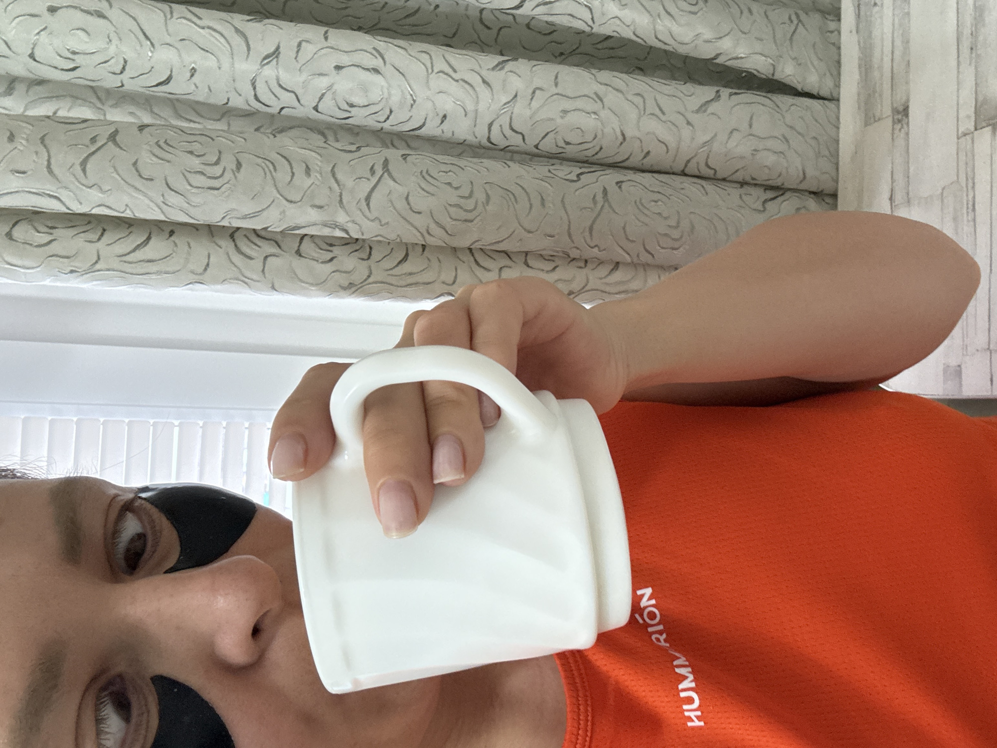 A selfie of a person with dark eye masks on, holding a white mug. The person is wearing an orange shirt with white lettering "HUMMRION" on the lower right. The background includes a patterned blind and a glimpse of window light. It is not possible to determine the location of Chaikovsky, Russia from the image.