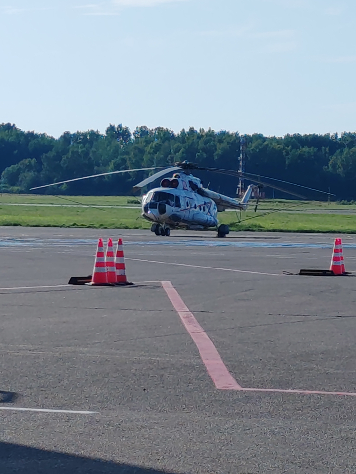 A large, white helicopter with blue and red markings is parked on an asphalt tarmac, likely at an airport. The helicopter has two main rotors and a tail rotor. It is viewed from the side, with its nose pointing to the left. The main landing gear is visible.
In the foreground, three orange and white striped traffic cones are placed in a line on the tarmac. A dark rectangular object is placed in front of the cones. A pink stripe painted on the tarmac runs from the bottom right corner upwards and to the left.
In the background, a line of lush green trees forms a dense forest. The sky above is a clear, light blue, suggesting a sunny day. The lighting indicates it is likely daytime, possibly late morning or early afternoon. There are no people visible in the image. The scene is quiet and still, suggesting the helicopter is either preparing for departure or has recently landed. There is no visible text in the image. The context provided indicates the location as Khrabrovo, Russia.