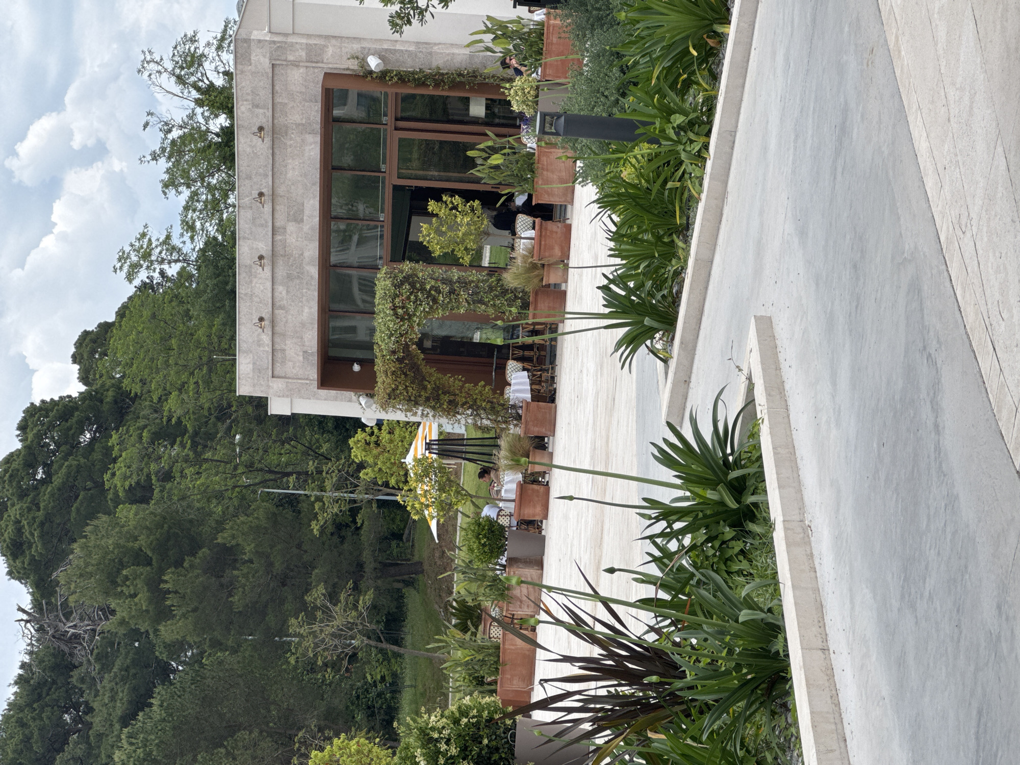 The image depicts an outdoor dining area in Tivat, Montenegro, situated adjacent to a modern, stone-clad building with large windows. The outdoor space features a tiled pathway lined with rectangular planters containing various plants, including leafy green shrubs and tall, spiky greenery. Tables with white linens and chairs are arranged along the pathway, suggesting a restaurant setting. People can be seen seated at some tables. In the background, lush green trees and vegetation provide a natural backdrop, and the sky is partially visible with scattered clouds. A low concrete wall runs along the edge of the pathway.