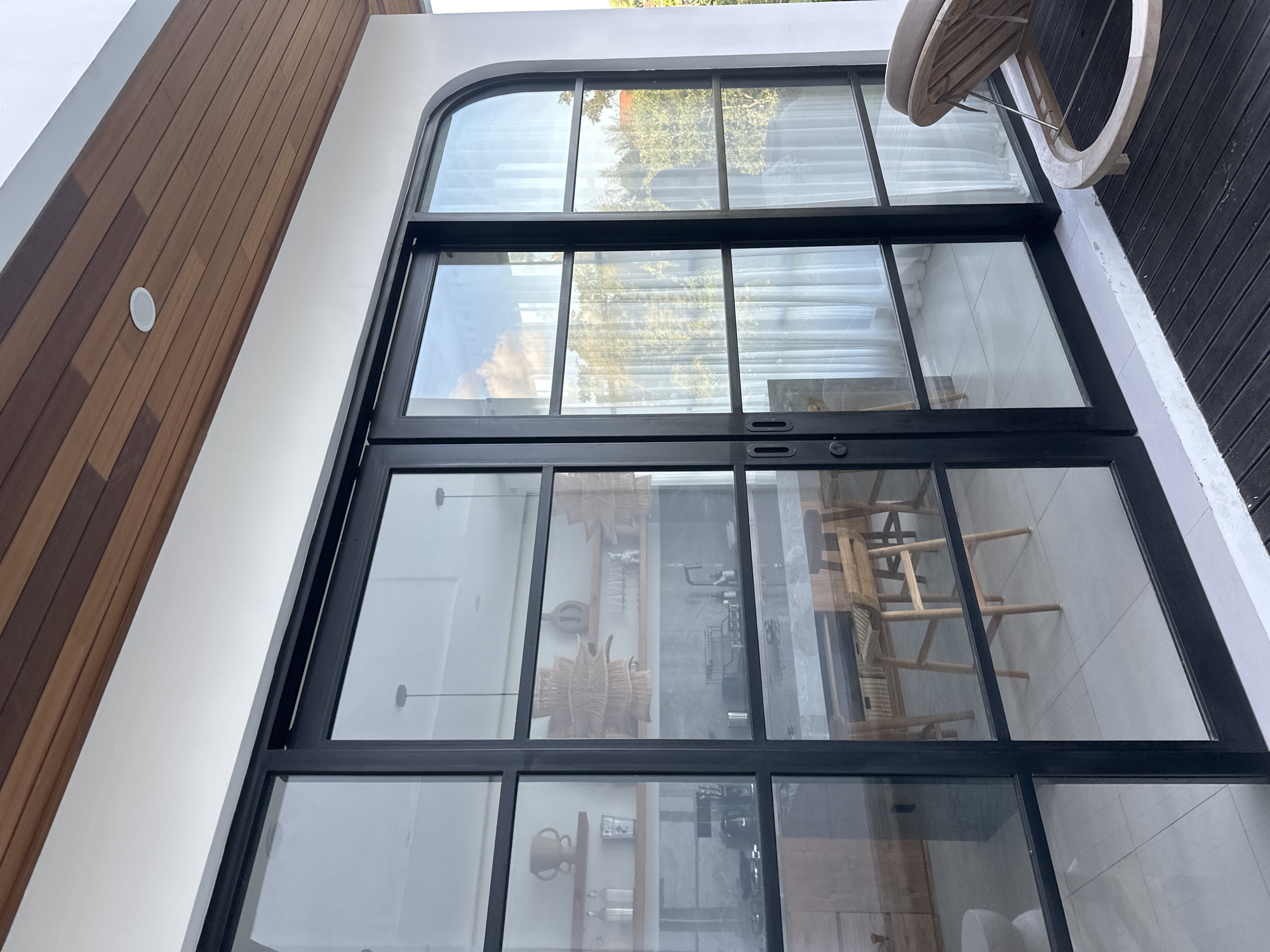 The image depicts a modern architectural exterior, likely a balcony or patio area, in North Kuta, Indonesia. The focal point is a large, black-framed, multi-pane glass door system with an arched top pane. This door system offers a view into an interior space that appears to be a kitchen or dining area, furnished with wooden chairs and decorative woven lamps. Reflections on the glass show the sky with clouds and trees, suggesting it's daytime.
To the left of the glass doors, a wall clad in horizontal wooden planks complements the white painted exterior wall. A circular white fixture, possibly a speaker or light, is embedded in the wooden cladding. To the right of the glass doors, a dark-colored deck or floor is visible, with part of a light-colored wooden chair or lounge visible at the top right corner of the image. The overall impression is one of contemporary design with natural materials. No people are visible, and the scene suggests a calm, possibly residential setting. The time of day appears to be daytime, and the weather is likely clear or partly cloudy, based on the sky reflections.