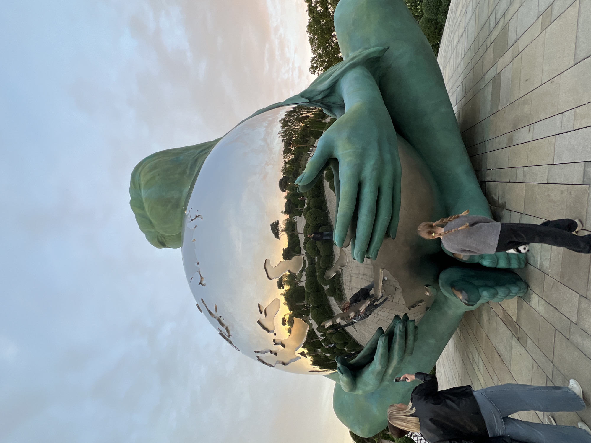 The image displays a large, green-patinated sculpture of a figure gently cradling a massive, highly reflective metallic sphere. The sculpture has a smooth, stylized human form, with prominent hands holding the gleaming orb.
Two individuals are present near the sculpture. One person, positioned to the right of the sphere (when viewed upright), wears a grey top, dark trousers, and has blonde hair in a braid, seemingly observing the reflections on the sphere's surface. The second individual, further to the left, is dressed in a black jacket and light blue jeans, also with blonde hair, and is holding a smartphone, likely capturing a photo or video of the artwork or the surroundings.
The metallic sphere acts as a mirror, reflecting the sky, trees, and other elements of the outdoor environment, including distorted images of people and distant foliage. The reflections show soft, warm light, suggesting late afternoon or early evening, possibly around sunset. The sky is a light blue-grey with subtle cloud formations.
The sculpture is situated on a paved surface, which extends to a textured stone or brick wall in the background, accompanied by green trees and well-maintained shrubbery. The setting appears to be an outdoor public space, such as a park or plaza, in Krasnodar, Russia. The scene captures an interactive moment with public art. There is no visible text in the image.