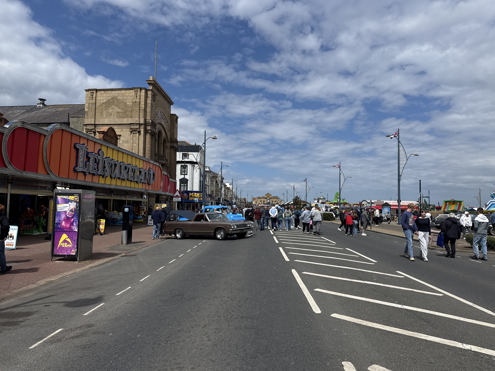 The image depicts a street scene in Great Yarmouth, United Kingdom, on a day with a mix of blue sky and scattered clouds. Along the left side of the street is a building with a large "Leisureland" sign. A brown Chevrolet El Camino is parked in the road, facing the right. A small crowd of people is gathered around the vehicle, many are dressed casually. The road is marked with white transverse stripes. Street lamps with Union Jack flags are visible along the right side of the road, near what appears to be a beachfront area with various stalls and amusements.