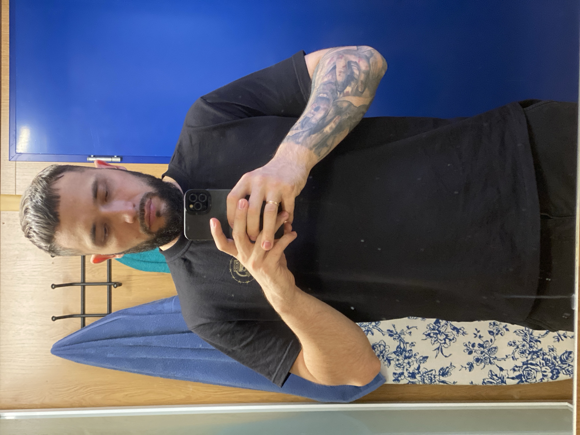 A man is depicted from the chest up, reflected in a mirror while taking a selfie. He has short, dark hair with graying strands on top, and a dark beard and mustache. His eyes are closed or looking downwards, creating a casual or contemplative expression. He wears a plain black t-shirt, featuring a faint, circular graphic on the right chest area that appears to include partially visible text, possibly starting with "RUBR..." or "RUBRIC," surrounded by a spiked or thorny design.
The man holds a dark-colored smartphone, likely an iPhone, in his right hand to capture the reflection. A simple gold wedding band is visible on his left ring finger. His left arm, fully visible on the right side of the image, is covered by a detailed sleeve tattoo in dark tones, seemingly depicting faces or figures.
The indoor setting suggests a bathroom or changing area. A bright blue wall or door serves as a prominent background element behind his head and to the left. To the right and below, light wooden paneling is visible. A black metal rack with hooks is partially seen behind his head. Below his torso, a folded blue towel or fabric is draped over an unseen object, and a smaller teal-colored towel is also present. The bottom of the reflection reveals a white fabric with a repeating blue floral pattern. No specific time of day or weather is evident. The location is Koskolovo, Russia.