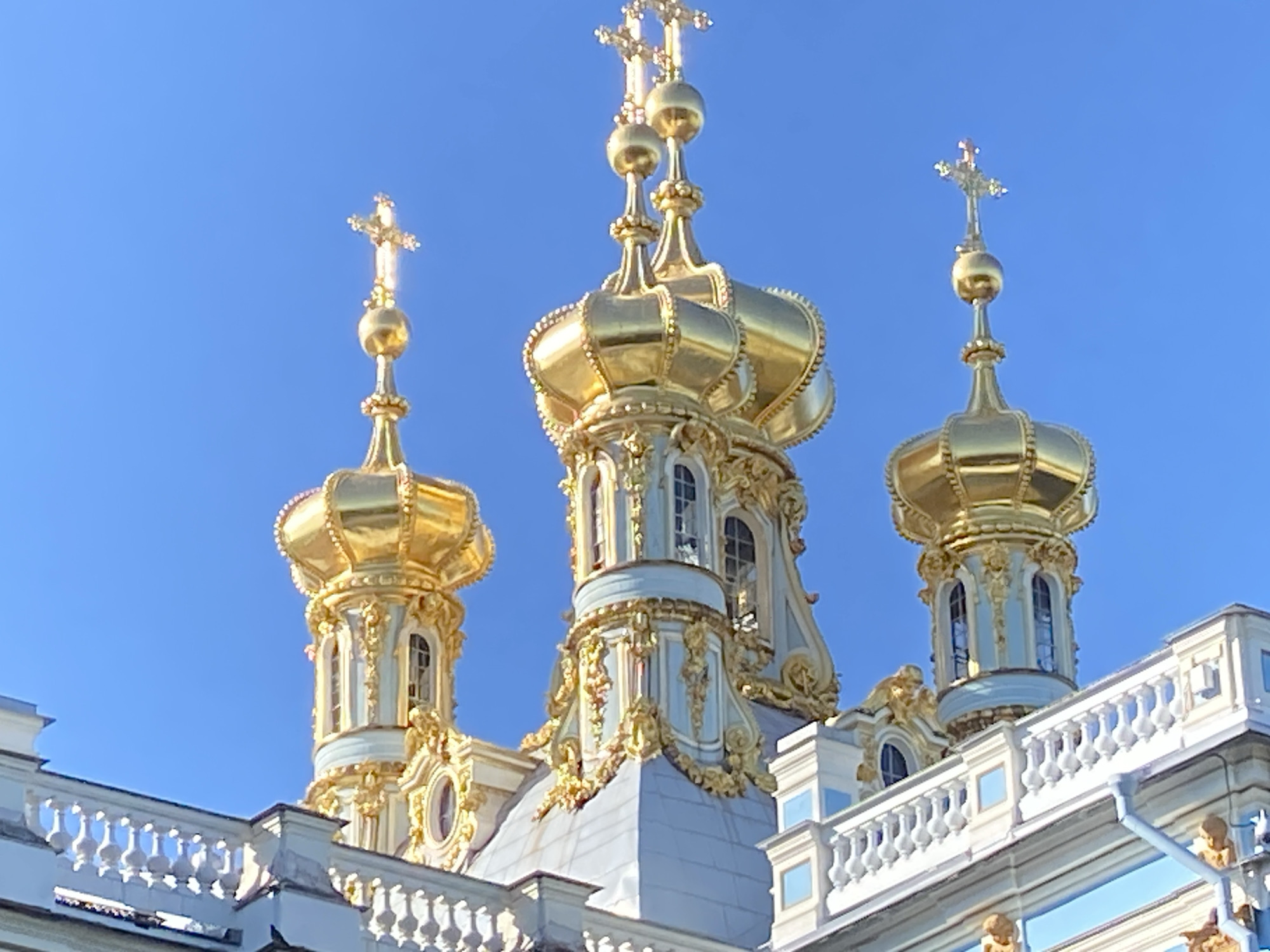 This is a medium shot, eye-level, outdoor, daytime photograph of the golden domes and spires of Catherine Palace in Pushkin, Russia, set against a clear blue sky. The ornate architectural details of the palace's roof are prominent, with multiple gilded domes topped with crosses. The domes are of varying sizes and shapes, with intricate carvings and decorative elements. The palace itself is a pale blue and white structure with white balustrades visible in the foreground. The lighting suggests a bright, sunny day, with strong sunlight reflecting off the gold surfaces.
