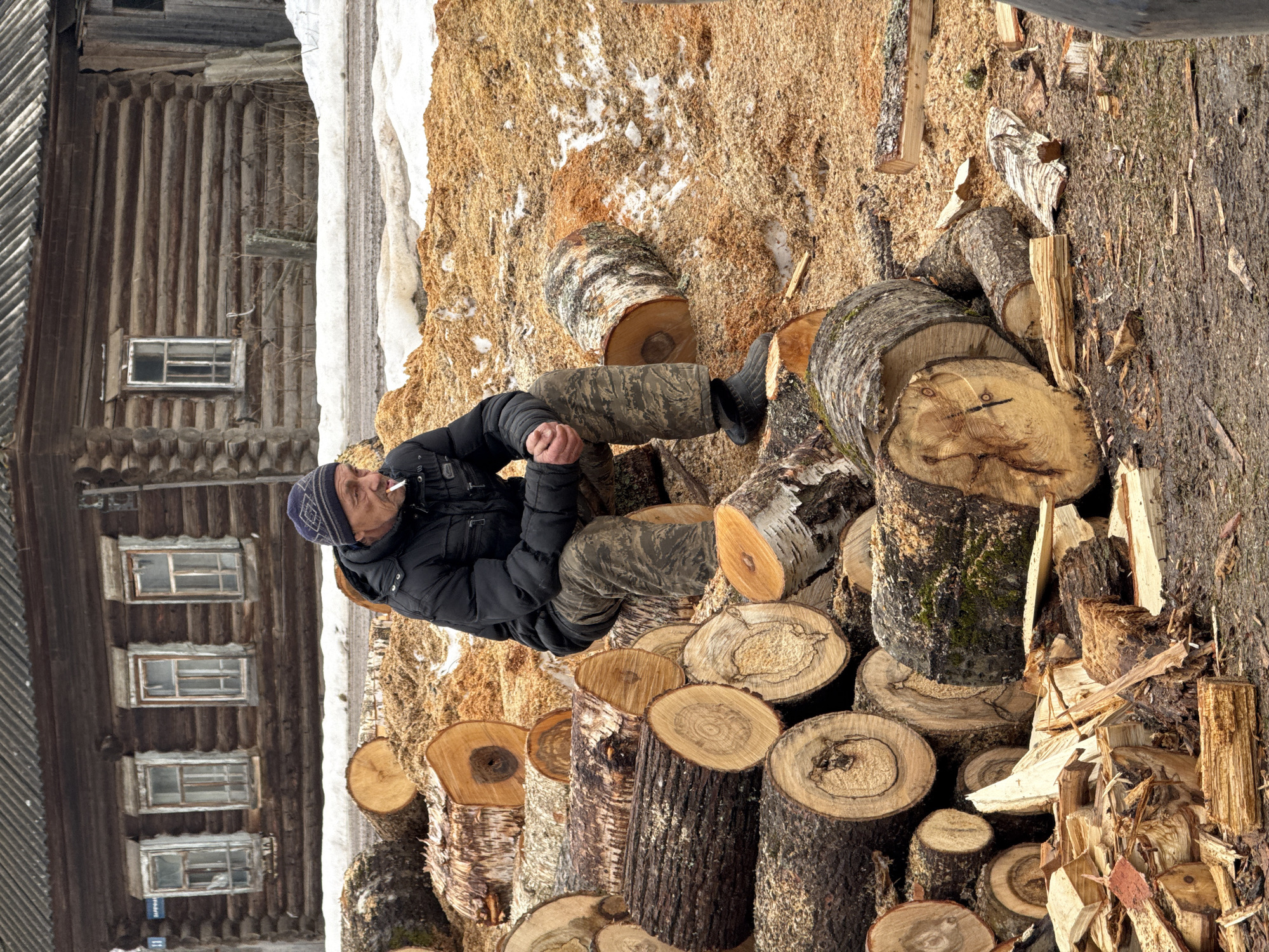 The image shows a man in a black jacket, camouflage pants, and a blue beanie sitting on a pile of freshly cut logs in Nizhnyaya Chermoda, Russia. He is smoking a cigarette. Behind him is a log cabin with four visible windows. The ground is covered in wood shavings and a patch of snow. Additional cut logs and chopped firewood are visible in the foreground.