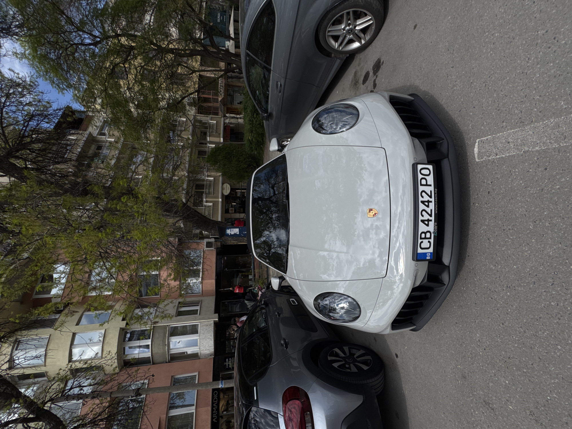 The image captures a daytime urban street scene in Plovdiv, Bulgaria, viewed from a slightly elevated and angled perspective. The main subject is a white Porsche sports car, likely a 911 model, parked on the right side of an asphalt road. Its front end, featuring classic circular headlights, a distinctive lower black grille, and the Porsche crest, is prominent.
The Porsche displays a Bulgarian license plate, "BG CB 4242 PO," with the text "www.t-cars.bg" visible on its holder. To the immediate left of the Porsche, a dark gray sedan is partially visible, showing its side profile and front wheel. Further back, the rear of another gray vehicle, possibly a hatchback or small SUV, is also partially seen.
The background features multi-story buildings, likely residential or commercial, with facades in varying shades of beige, light yellow, and reddish-brown. Numerous windows, some reflecting the sky, punctuate the building exteriors. Lush green trees with slender branches and dense foliage line the street, partially obscuring the architecture and revealing patches of clear blue sky above. Visible text on building signs includes "ARMADIO" and a partially visible "BALMAIN" (inverted). The road surface is asphalt with faint white markings. The scene conveys a typical, calm urban setting.