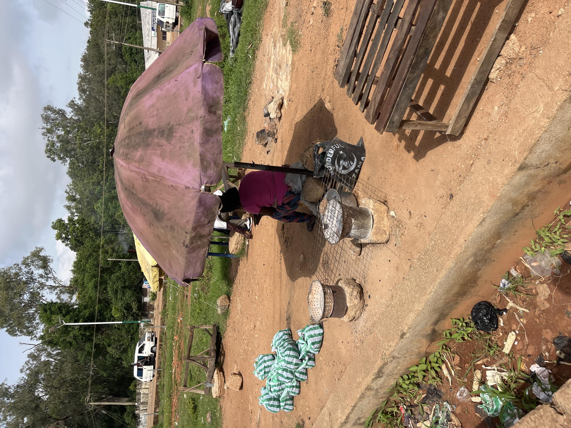 In Bukuru, Nigeria, a person sits under a large, weathered pink umbrella, shaded from the sun. They are wearing a pink shirt and patterned pants. Several green and white striped bags are neatly arranged on the ground to the left of the umbrella. Two metal cooking stands, each resting on a rock, are positioned in front of the umbrella. A black bag with the word "WONDERFUL" printed on it lies near the seated person. To the right, a simple wooden bench is placed on the reddish-brown dirt ground. In the background, trees line a street where a white van is visible. The scene is set outdoors on a cloudy day.