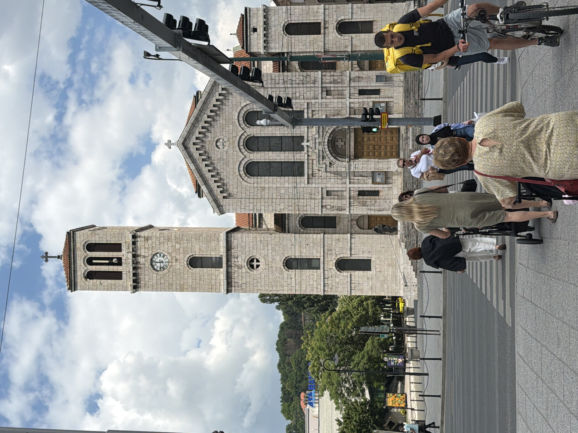 The image shows a street scene in front of the Sacred Heart Cathedral in Sarajevo, Bosnia and Herzegovina. A light-colored stone cathedral with a tall bell tower is the central architectural feature. A cross is visible at the apex of the bell tower and the main facade. Several people are crossing a pedestrian street in the foreground. One person is riding a bicycle with a bright yellow delivery backpack. Other individuals are walking, with some pushing strollers. Traffic lights and street lamps are also visible. In the background, there are trees and buildings, and the sky is partly cloudy.