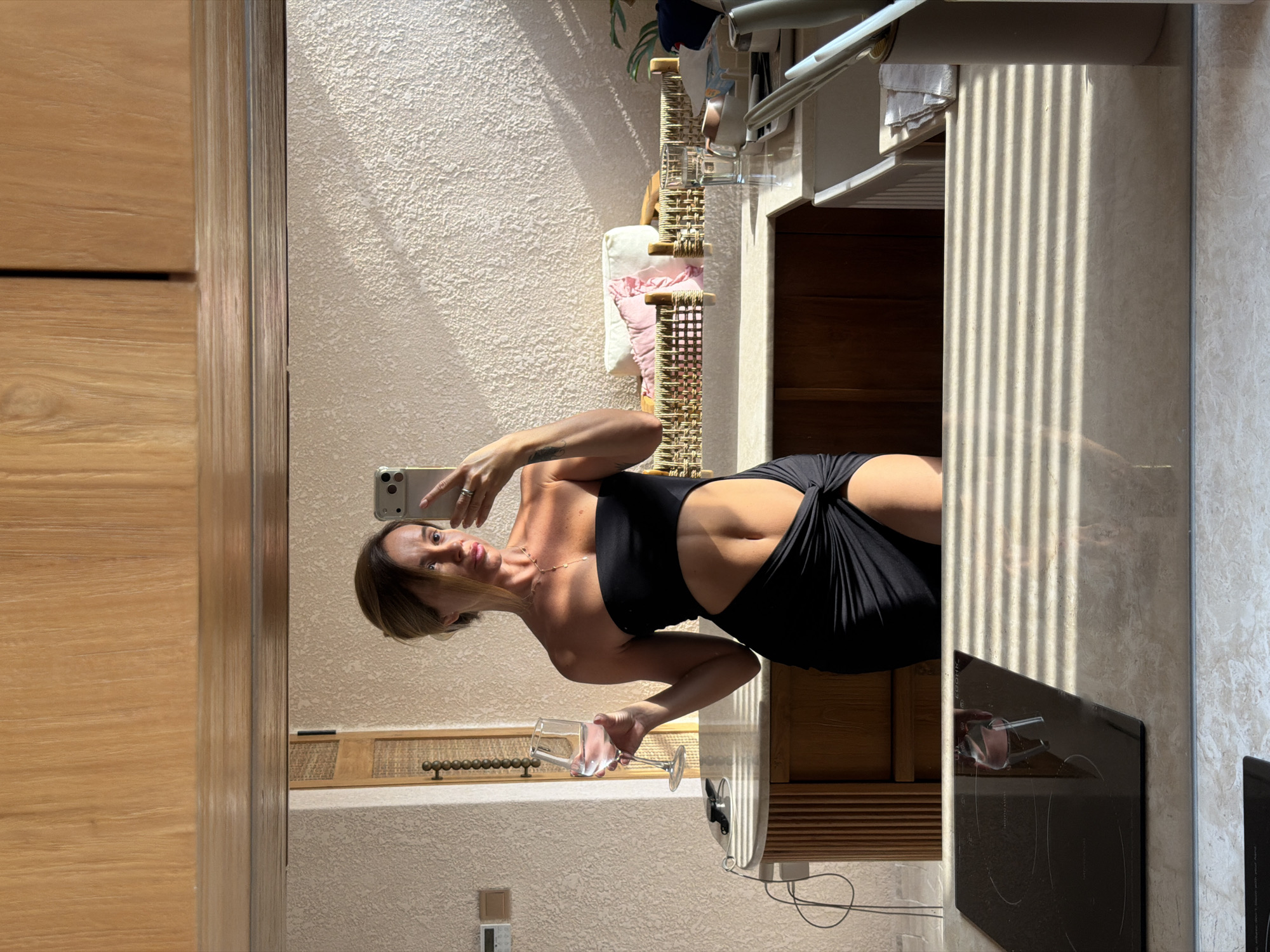 A woman, viewed in a mirror reflection, stands in what appears to be a kitchen or bathroom area. She is holding a wine glass in her left hand and a smartphone in her right hand, seemingly taking a selfie. She is wearing a black, strapless, cut-out dress. The background shows wooden cabinetry, a textured wall, and various items on a countertop, including what looks like toiletries and a sink. The overall lighting suggests it is daytime, with sunlight casting shadows. The location is identified as Mengwi, Indonesia.