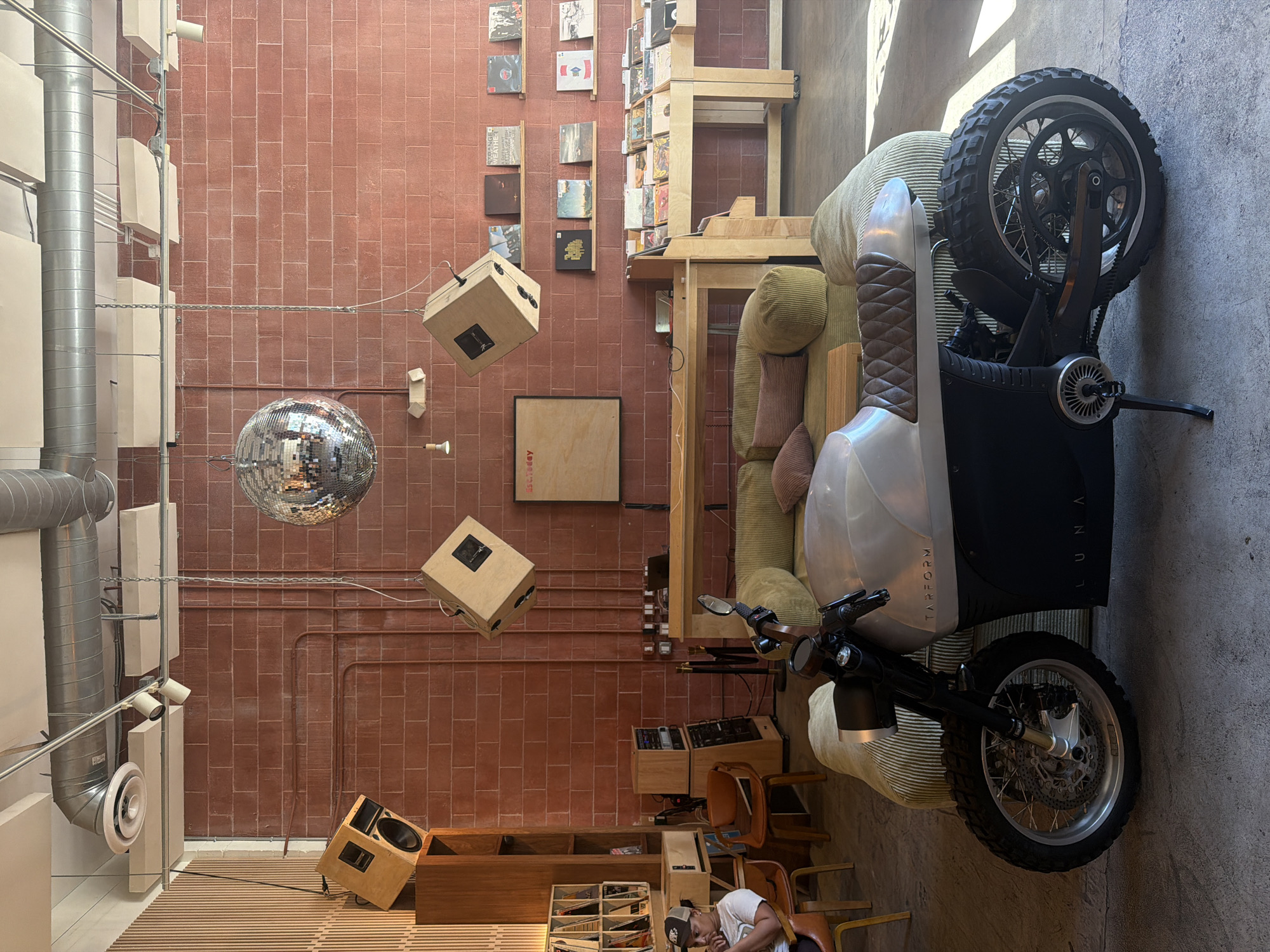 A tilted interior view in Los Angeles, United States, showcases a unique blend of design and functionality. The foreground features an electric motorcycle parked sideways on a light green couch. A person is seated in a brown chair near a wooden cabinet filled with media. The midground displays a brick wall adorned with vinyl records, interspersed with box-shaped speakers suspended by chains. A disco ball hangs from the ceiling above the speakers. The background includes a ventilation system and a window providing natural light.
