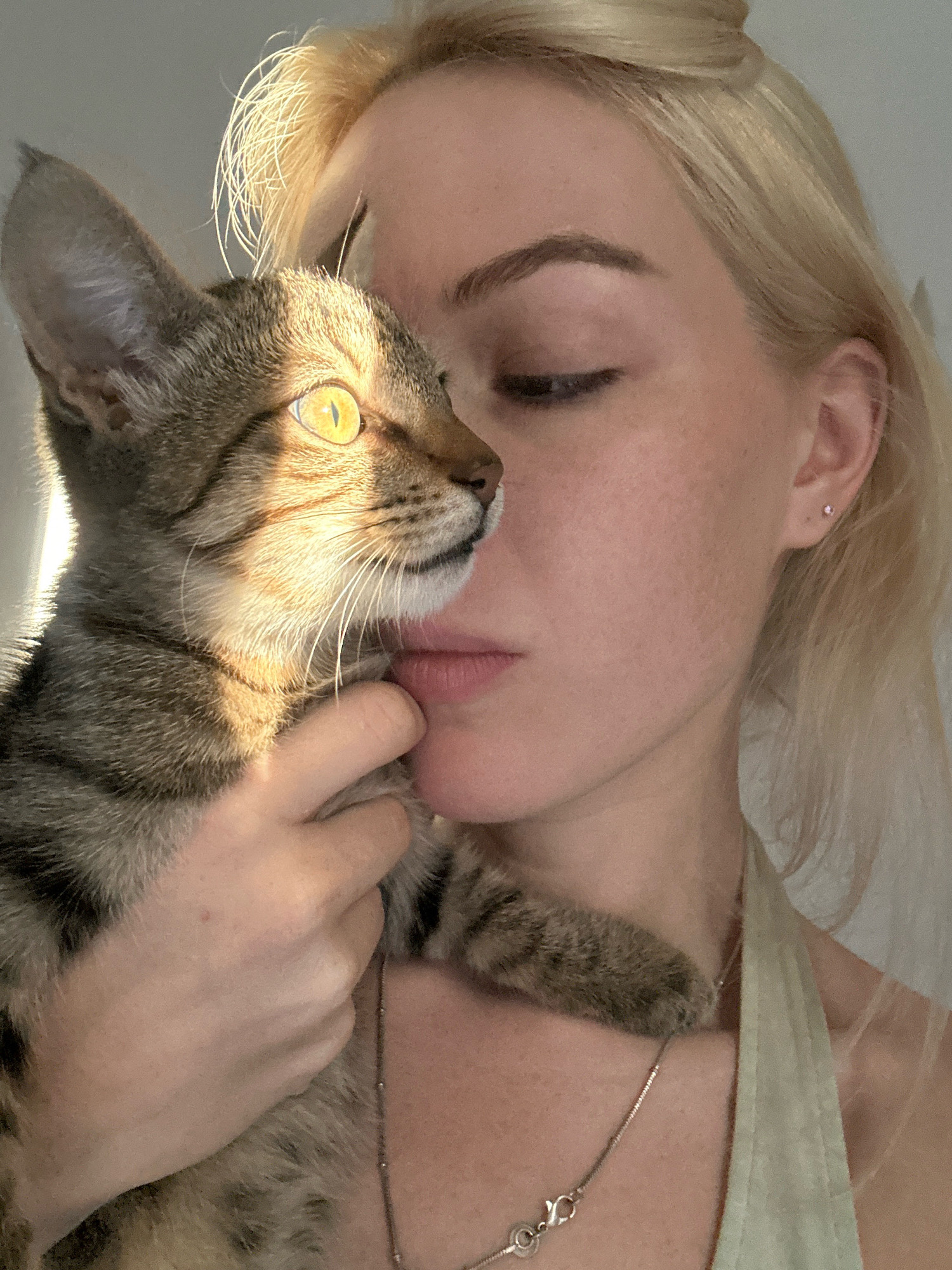 This is a close-up, candid photograph of a young blonde woman holding a tabby kitten. The woman's eyes are closed, and she is leaning her head towards the kitten, who is looking to the left with bright yellow eyes. The kitten's fur is highlighted by warm sunlight, creating a dramatic effect on its face and ears. The woman is wearing a light green tank top and a silver necklace. The background is a plain, light-colored wall. The image appears to have been taken indoors, possibly in a residential setting in Tbilisi, Georgia, given the lighting and the intimate nature of the pose.