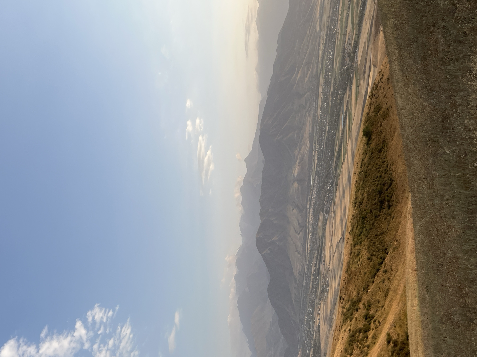 This image captures a serene, expansive view of the outskirts of Besh Kungei, Kyrgyzstan. The foreground is dominated by a gently sloping, dry grassy hillside, casting a long shadow across the land. Behind it, the terrain transitions to golden, cultivated fields, interspersed with darker green patches, suggesting agricultural activity. A small village or town is visible nestled in the valley below, characterized by its clustered buildings and roads. The mid-ground is defined by the layered, muted tones of imposing mountains that recede into the hazy distance. The sky above is a clear, pale blue, with a few wispy white clouds scattered across the upper left quadrant, hinting at late afternoon or early evening light. The overall scene is one of natural beauty and rural tranquility, with a human settlement integrated into the vast landscape.