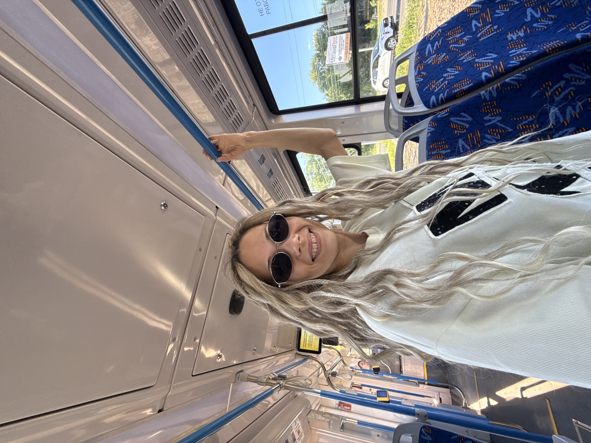 The image depicts the interior of a public transit vehicle, likely a bus or trolleybus, in Rusanovo, Russia. A woman with long, blonde dreadlocks, dark oval sunglasses, and a light-colored (off-white) long-sleeved top stands in the foreground, smiling towards the camera. Her right hand grips a blue horizontal handrail located above her head.
The vehicle's interior features a white ceiling with integrated lighting strips and air vents. Blue horizontal and vertical handrails are visible throughout the cabin. To the right, two empty blue fabric seats with a distinctive patterned design (orange, light blue, and dark blue geometric shapes) are visible. On the left side, a yellow digital display screen with Cyrillic text and yellow request buttons on vertical poles are present.
Through the large windows on the right, a clear blue sky, green trees, and a paved road are visible. A white SUV, possibly a Haval Jolion, is seen on the road. A white rectangular sign with the prominent Cyrillic text "МеталлПроф" (MetallProf) is visible outdoors, along with other partially obscured Cyrillic text on the upper right window. The vehicle's floor is a dark surface reflecting light.