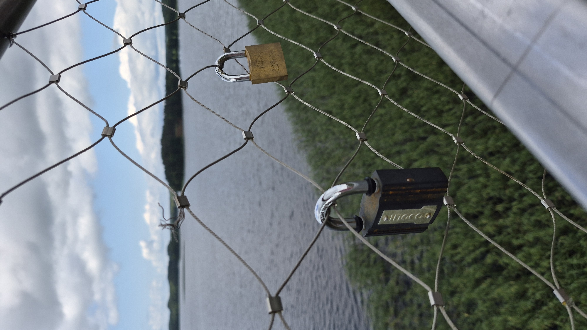 A close-up, low-angle shot captures a wire mesh fence with two padlocks attached. The padlock on the right is brass and has faded writing on it, including the numbers "2021/06/2". The padlock on the left is dark gray and black, with some rust and a visible label. The fence is made of thin, interwoven wires held together by small square connectors.
Beyond the fence, a body of water stretches to the horizon, with a treeline visible in the distance. The water is a muted gray-blue, and the sky above is partly cloudy with white clouds against a blue sky. Dense green reeds or tall grass line the near edge of the water.
The image was taken in Zarasai, Lithuania, likely on a bridge or viewing platform overlooking Lake Zarasas, a popular spot for couples to attach "love locks."