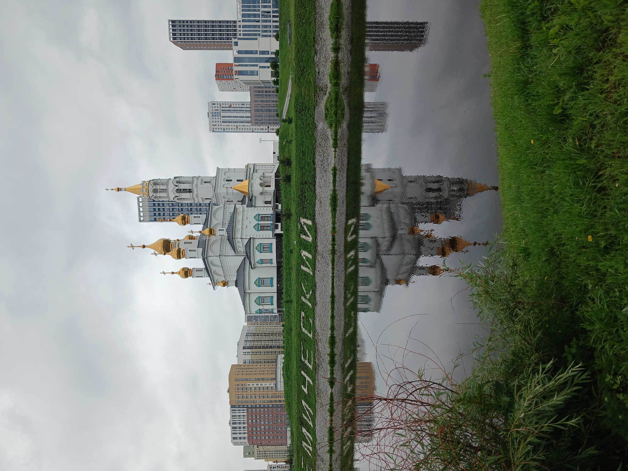 The image captures a scene in Yekaterinburg, Russia, featuring the Holy Trinity Cathedral and a modern cityscape. The cathedral, with its white walls and golden domes, stands prominently on a grassy hill. Behind it, a cluster of tall, modern buildings with glass facades rises against a cloudy sky. In the foreground, a body of water, likely a river, reflects the cathedral and the surrounding buildings, creating a symmetrical mirror image. Green grass covers the banks of the water and the hillside. In white letters on the riverbank, the word "РОССИЙСКИЙ" (Russian) is visible. No people are visible in the image. The overall atmosphere is overcast and tranquil.