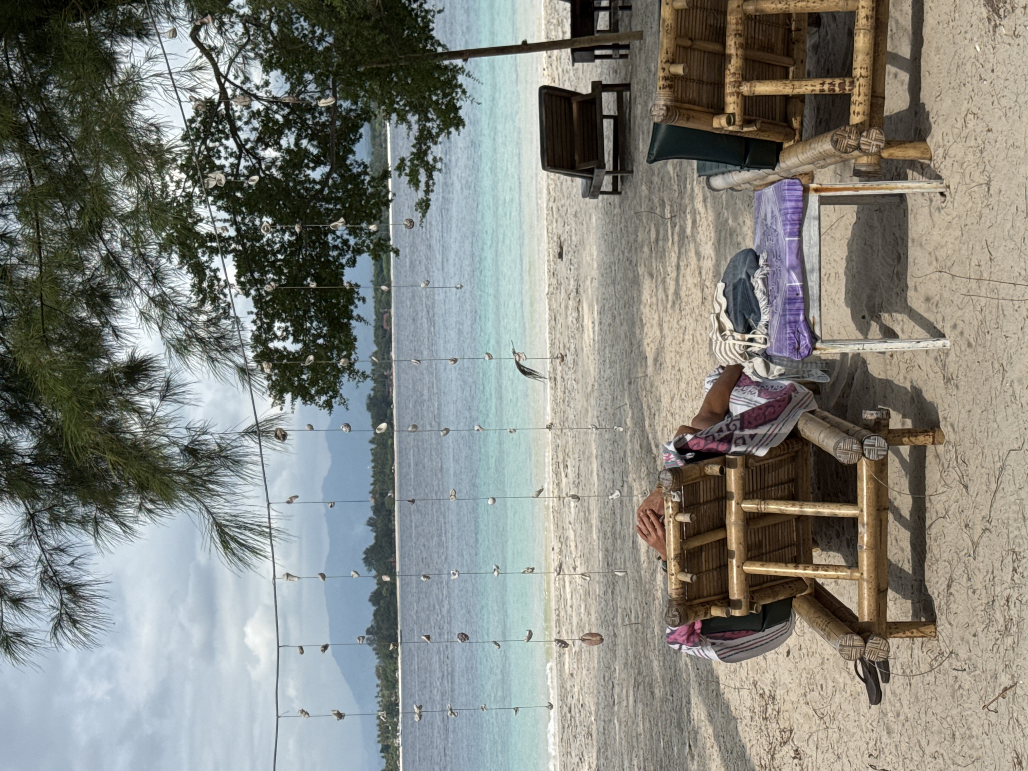A beach scene in Unknown, Indonesia. The sand is light-colored and there is a light blue sea with trees on the far shore. There are bamboo structures on the beach, including chairs, and hanging strings of shells. A person's hands can be seen sitting on a bamboo chair.