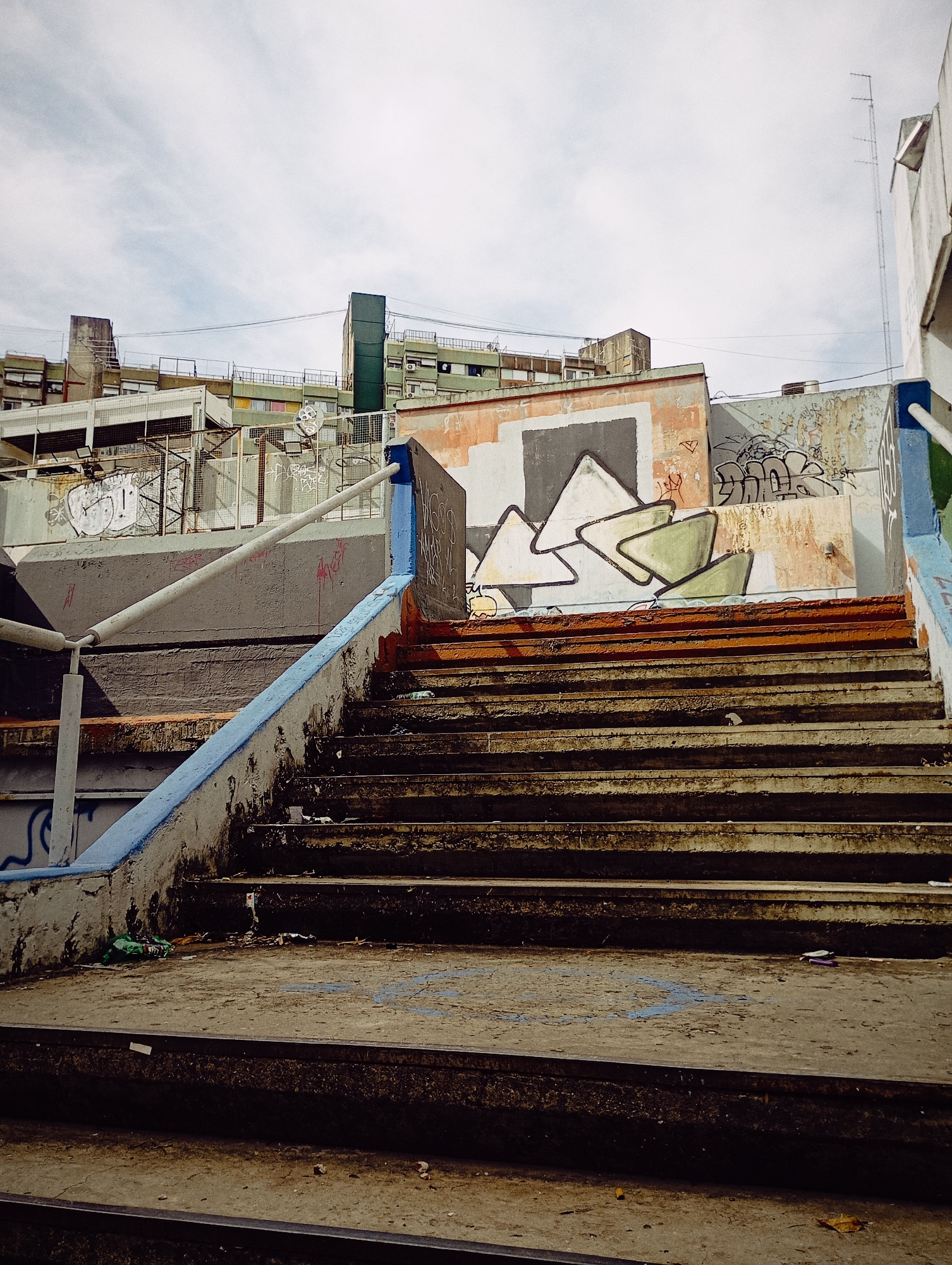 A set of outdoor concrete stairs with blue railings is depicted, leading up to a concrete wall adorned with graffiti. The stairs show signs of wear and dirt, with trash scattered on some steps. The wall behind the stairs features various graffiti tags, including abstract geometric shapes and illegible writings. The building complex in the background appears to be an urban residential area, composed of multiple buildings of varying heights. The sky is partly cloudy, casting an even light across the scene. The location is Buenos Aires, Argentina, as identifiable by distinctive graffiti styles and architecture.