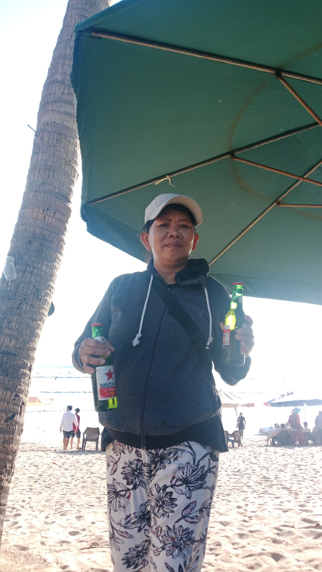 A woman wearing a gray zip-up hoodie, white baseball cap, and floral patterned pants stands on a sandy beach, holding two glass bottles. The bottle in her left hand is partially visible and has a red and white label with a star. The bottle in her right hand is green and appears to be illuminated by sunlight. Behind her, a palm tree is on the left, and a large, green umbrella with metal supports is overhead. In the background, other beachgoers are visible, along with beach chairs and umbrellas, and the ocean. The overall lighting suggests a sunny day.