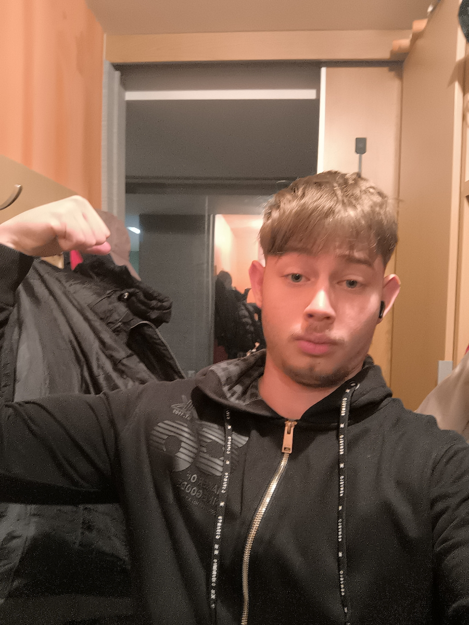 A young man with light brown hair poses in what appears to be an interior hallway or entrance area. He is wearing a black hooded zip-up jacket, and flexing his left arm. Hanging beside him is a black jacket. In the background, there's a doorway leading to a dimly lit space and adjacent to that is a wall. Based on the style of architecture and general appearance, there is no indication of this being in Sofia, Bulgaria.