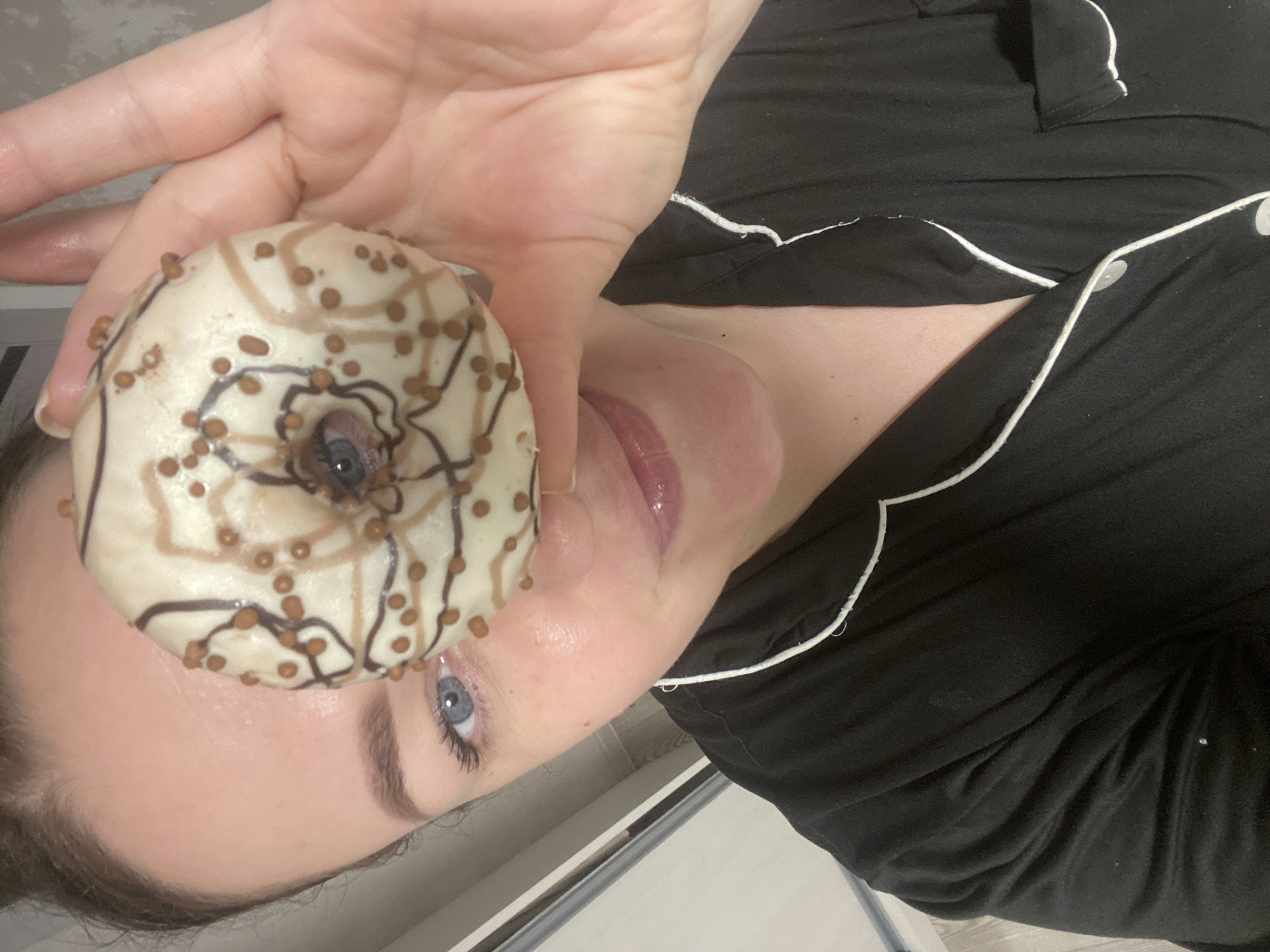 The image depicts a close-up, high-angle view of a female individual indoors in Sochi, Russia. She holds a ring-shaped donut with her right hand, positioning it directly in front of her face. The donut is covered in white icing, decorated with irregular dark brown wavy lines and numerous small, light brown spherical sprinkles. Her left blue eye is visible through the donut's central hole. Other partially visible facial features include her forehead, left eyebrow, nose, and glossy lips, which show a slight upturn. She wears a black garment with white piping, consistent with pajamas or loungewear, visible on her left shoulder and chest. The background consists of a light-colored surface, with a vertical light gray metallic strip visible on the far left.