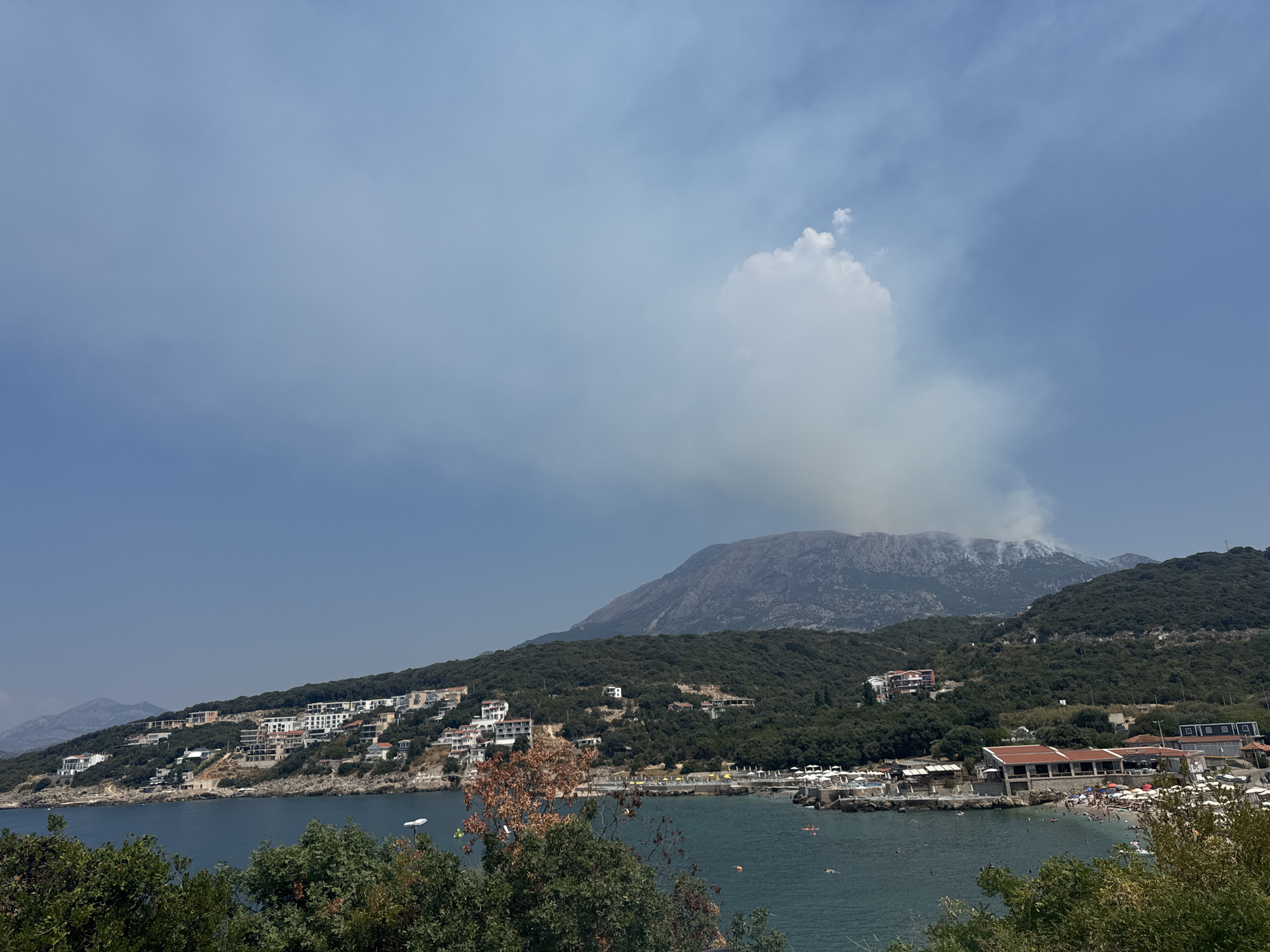 This is an eye-level, wide shot captured on a bright, hazy day in Kunje, Montenegro. The scene overlooks a tranquil bay where the dark blue water is dotted with swimmers and a few small boats. A sandy beach, adorned with numerous white umbrellas and loungers, lines the shore. Buildings, some with terracotta roofs, are nestled into the lush, green hillside that slopes down to the water. In the background, a large, imposing mountain dominates the landscape, with a plume of smoke rising from its peak, suggesting a wildfire. The sky above is a pale blue, partly obscured by scattered clouds and the haze.