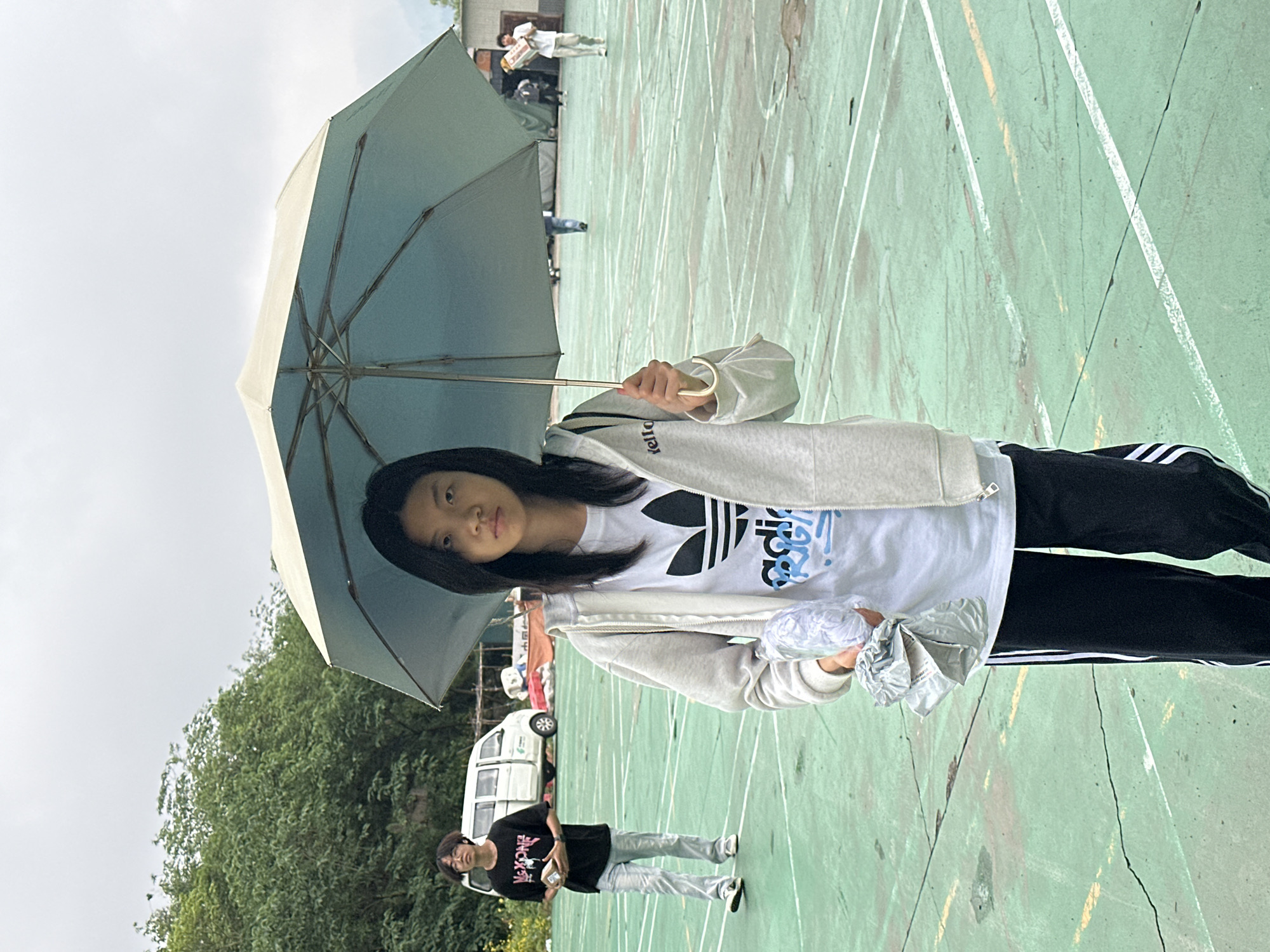 The image shows a young woman holding an umbrella on what appears to be an outdoor sports court. The surface is green with white lines marking the boundaries. The woman is wearing a white t-shirt with a black Adidas logo, a light grey hoodie, and black pants with white stripes down the side. She is also holding a crinkled plastic bag. In the background, there's another person walking and a white van parked. The surrounding environment includes trees and buildings, suggesting an urban or suburban setting. Given the reference to Jiangjin, China, it is likely that the photo was taken in or near that city.