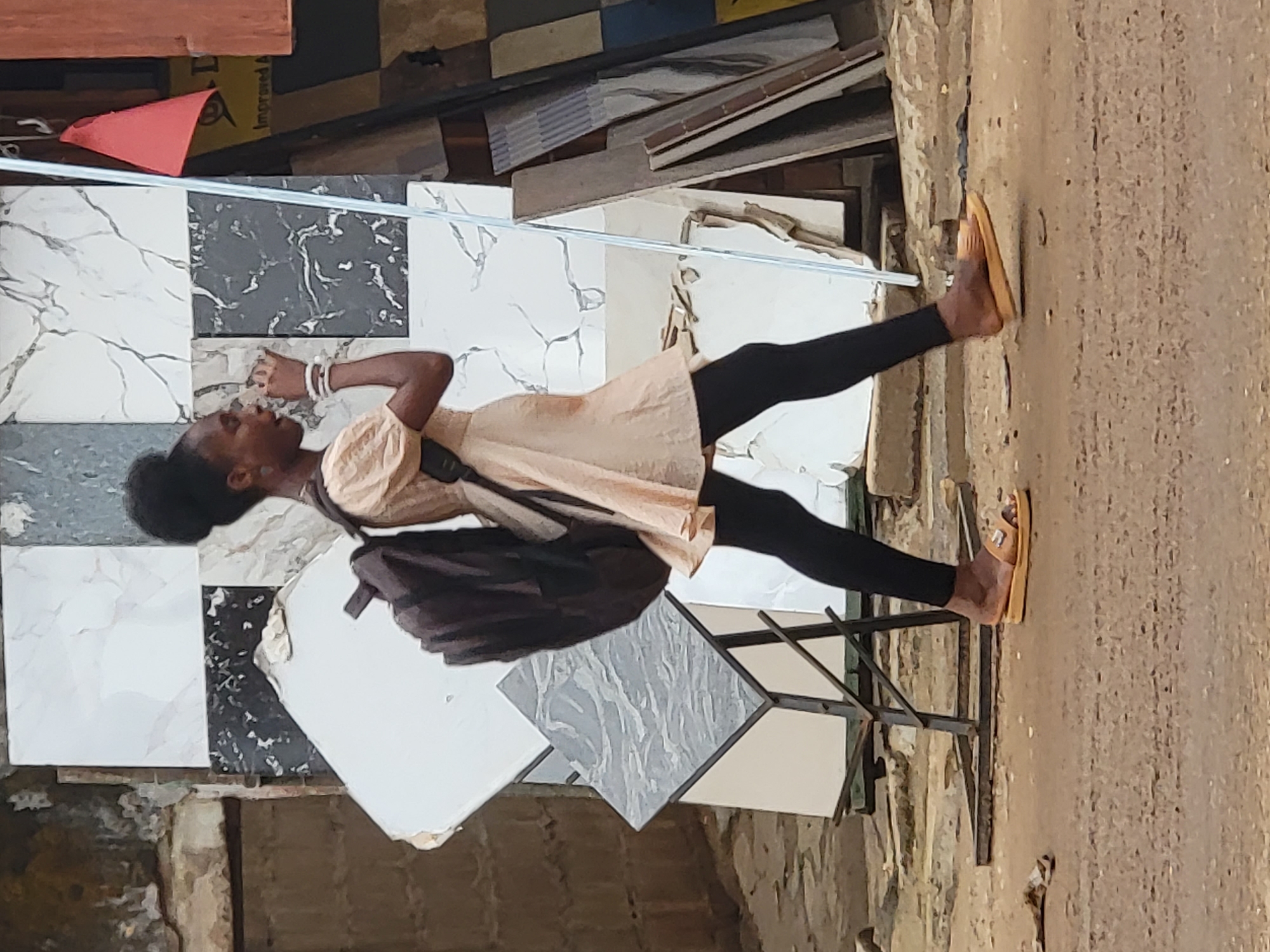 A young person stands in what appears to be an outdoor market or building materials store in Maiburiji, Nigeria. The individual has dark skin, with black hair pulled back, and is wearing a light-colored tunic or dress over black leggings, paired with light brown open-toed sandals. A dark shoulder bag is visible on their right side, and white bracelets are on their left wrist. Their left hand is slightly raised, and their mouth is slightly open, suggesting they are speaking or actively observing something.
They are standing in front of and around a display of large square tiles, predominantly white with grey marbling, black with white marbling, and some light grey patterned tiles. These tiles are arranged on a dark metal stand and leaning against surfaces. A thin, light-colored rod or pipe runs horizontally across the mid-ground.
In the background, stacks of what appear to be wooden planks or other building materials are visible. A red, triangular object is in the upper left background. The ground is an unpaved, textured dirt or earth surface. The scene is brightly lit by daylight, indicating clear weather. On a yellow surface in the upper background, faint text reading "Improved A" or "Improved N" along with a triangular symbol can be observed. The overall setting suggests a commercial area focused on construction or home improvement supplies.