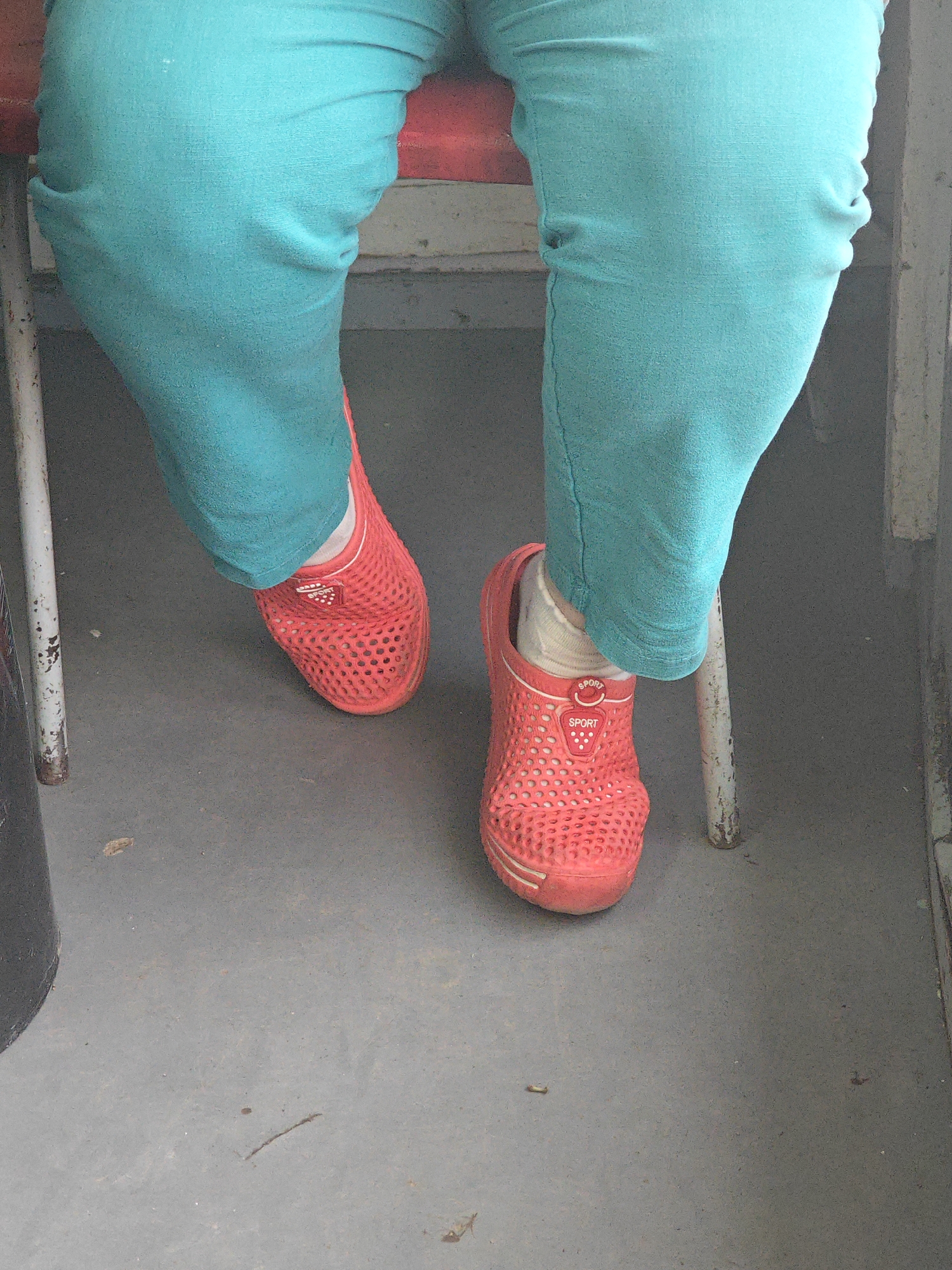 The image shows the lower body of a person wearing teal pants and red perforated clogs. The person is sitting on a red and metal chair, with their feet resting on a gray floor. The scene is likely indoors, possibly in a public space like a bus or waiting room, given the utilitarian furniture and floor. The background is blurred, but appears to be a neutral-colored wall. There are no distinguishing features that would pinpoint the exact location within Krasnoyarsk, Russia, but the overall setting suggests a mundane, everyday environment. The focus is entirely on the person's footwear and attire.