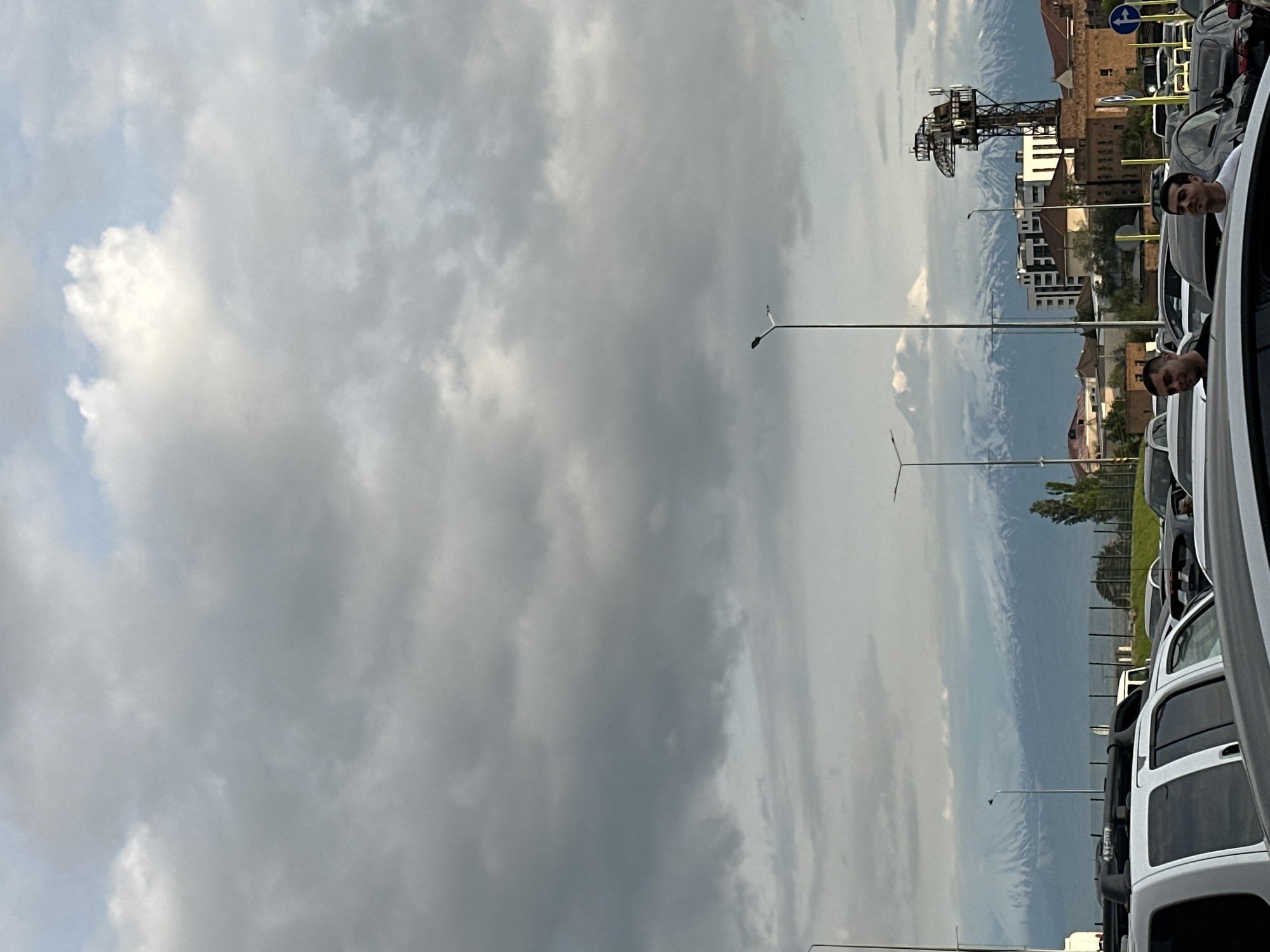 A photograph captures a Yerevan, Armenia street scene filled with parked cars. The sky is predominantly cloudy. Two men are visible, peering from car windows. Streetlights line the street. Buildings, including a structure resembling a tower, and mountain ranges are in the background.