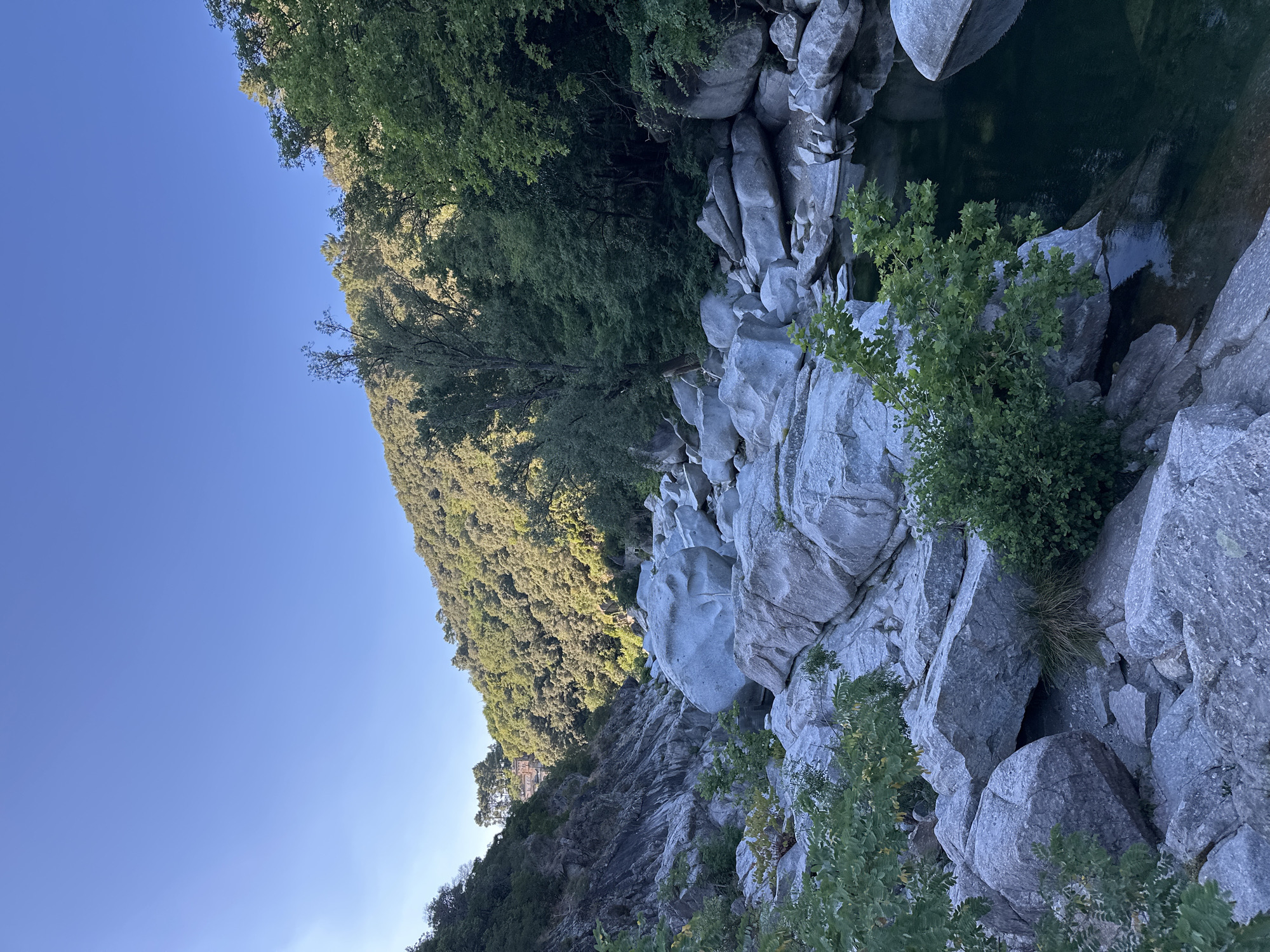 The image depicts a scenic view of the Gardon River gorge near Anduze, France. The gorge is characterized by large, smooth, gray boulders that have been sculpted by the river over time. Lush green vegetation, including trees and bushes, grows on the slopes of the gorge and among the rocks. The water in the river is dark and appears calm in places. The sky above is a clear, bright blue, suggesting a sunny day. In the distance, on the upper slope of the gorge, a building with a reddish-brown facade and a tile roof is visible, nestled amongst the trees. The overall impression is one of natural beauty and tranquility.