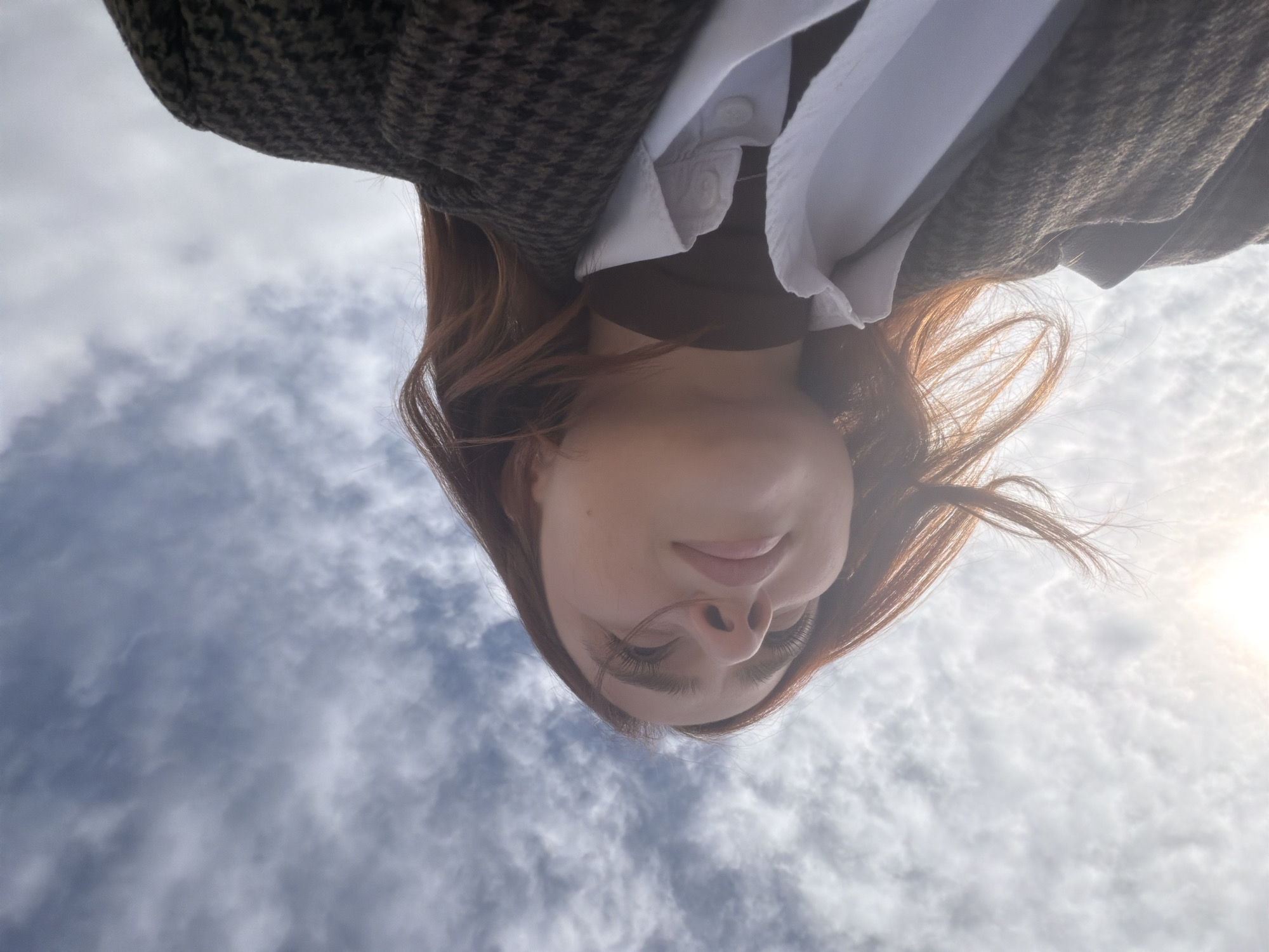 In Les Trois-Domaines, France, a low-angle outdoor shot captures the upper torso and head of a female individual looking upwards against a cloudy sky. The subject has reddish-brown hair, some strands of which are visibly windblown, and prominent eyelashes. She is dressed in a dark, patterned outer coat, worn over a white collared shirt that appears unbuttoned at the top, and an underlying dark, high-neck garment. The sky is partially obscured by scattered white and grey clouds, with a bright light source, consistent with the sun, visible in the upper left, causing a lens flare effect. No other individuals or distinguishing urban features are present.