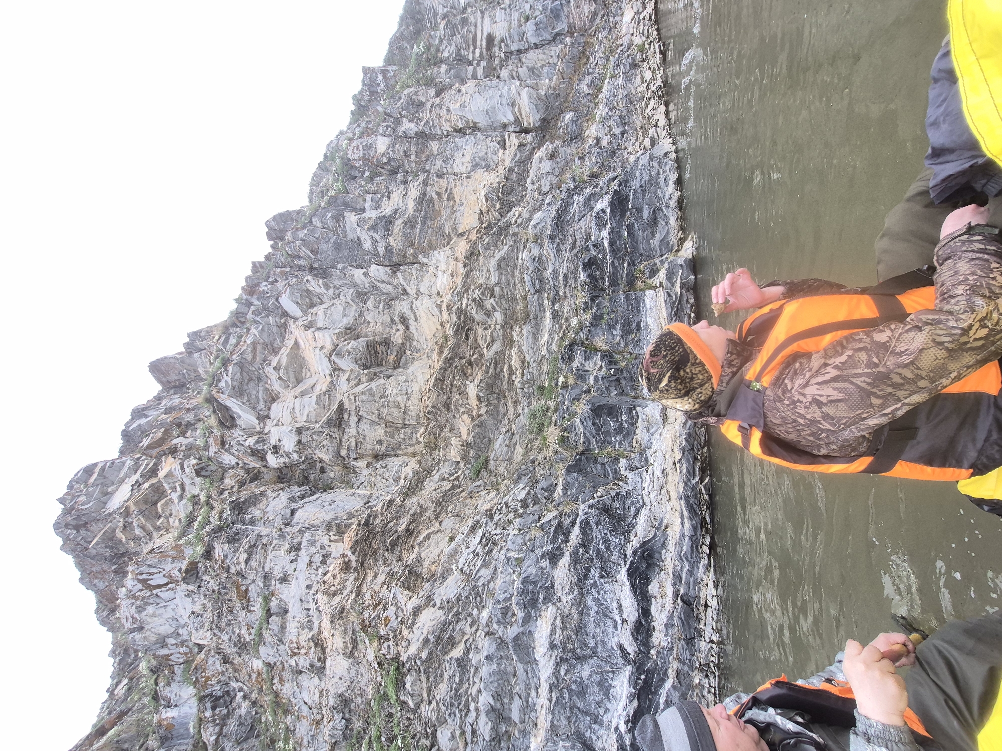 The image captures a scene from a boat or raft on a body of water, positioned alongside a towering, rugged rock cliff. In the foreground, on the right side of the frame, a person is visible from the chest up, wearing an orange life vest over a camouflage-patterned long-sleeved shirt and a matching camouflage hat or hood. This individual appears to be looking upwards towards the cliff face. Partially visible in the upper right, another person's lower body is seen, with legs clad in green trousers, and a hand with a watch on the wrist, possibly holding an object.
The left and central portions of the image are dominated by the massive rock cliff, which rises steeply from the water. The cliff face is highly textured, displaying a variety of grey, black, brown, and white tones, suggesting different rock strata and weathering. Small patches of green vegetation cling to crevices and ledges. The water itself, filling the foreground and extending towards the cliff, is murky green-brown with subtle ripples, reflecting the ambient light. Above the cliff, in the upper left, a bright, overcast white sky is visible.
The scene depicts an outdoor water activity, likely rafting or boating, given the presence of life vests and the perspective from the water level. The time of day appears to be daytime, characterized by bright, diffused lighting, indicative of an overcast sky or soft natural light. There is no visible text in the image. The location is Novomunasipovo, Russia.