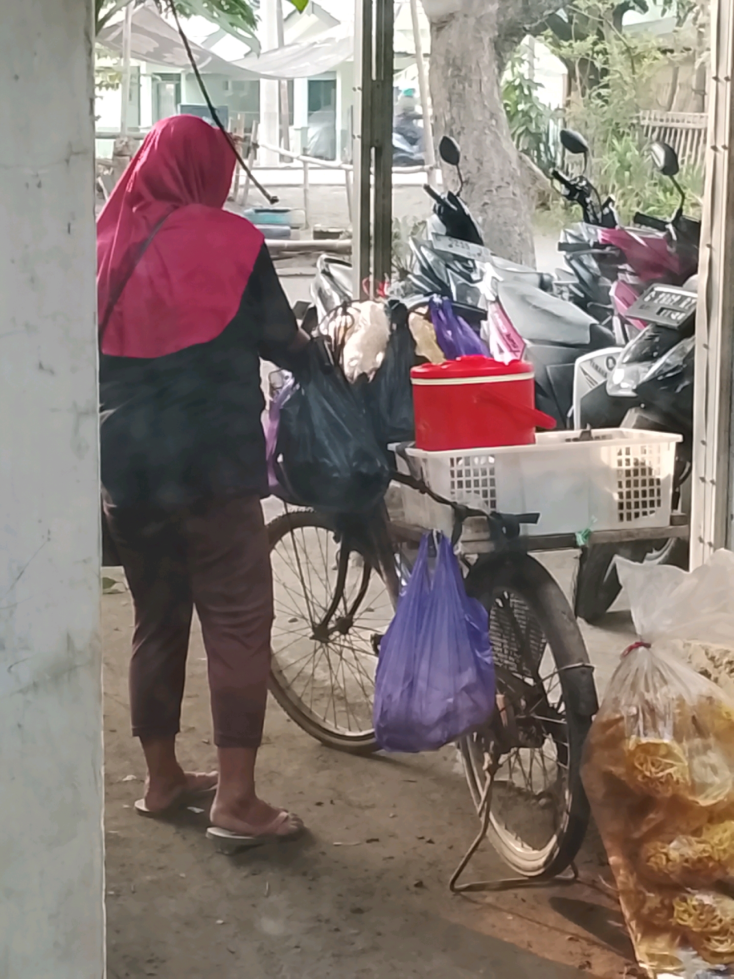 An over-the-shoulder view of a person in a red hijab and a dark shirt is seen from the back, standing beside a bicycle laden with various items in Ujungrusi, Indonesia. The person is wearing dark trousers and light-colored sandals. The bicycle has a white basket on its rear rack, containing a red insulated container and other objects. Two purple plastic bags hang from the bicycle, one from the handlebars and another from the rear. To the right of the bicycle, a large clear plastic bag filled with yellow, puffed snacks is visible. In the background, several motorcycles are parked, and a building with light-colored walls and windows can be seen. The ground is a dirt or gravel surface.
