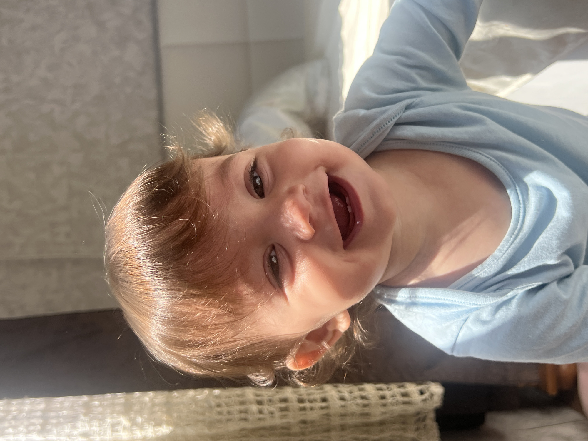 The image shows a baby wearing a light blue shirt lying on a surface with a white crocheted blanket in the foreground. The baby is facing the camera with an open mouth, indicating a smile or laughter. The baby has brown hair and eyes, and fair skin. Sunlight casts shadows across the scene, suggesting a daytime setting. The background is blurred, but it appears to be an indoor setting with a wall. There is no information to suggest that the image was taken in Elena, Bulgaria, or any specific location.