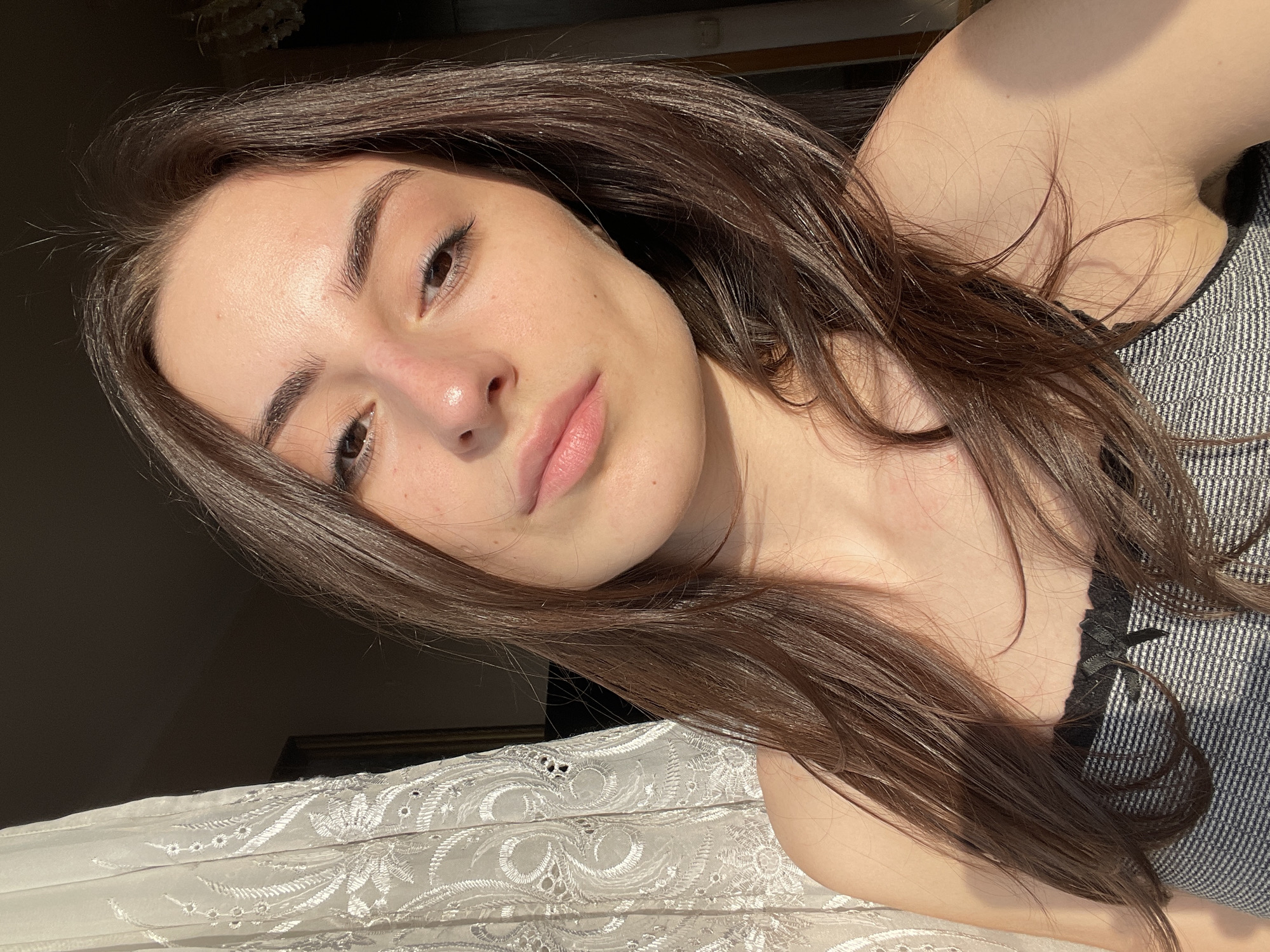 A young adult female is depicted in a close-up, self-portrait style image captured indoors. She possesses long, dark brown hair, parted centrally, extending over her shoulders. Her complexion is fair, with visible freckles across her cheeks and nose. Her eyes are dark brown, and her gaze is directed towards the camera. Her lips are a natural pink hue, slightly parted. She is wearing a sleeveless top or camisole featuring a horizontal grey and white striped pattern, trimmed with black lace and a small black bow centered at the bust line.
Strong, directional sunlight illuminates the right side of her face and body (viewer's left), casting pronounced shadows on her left side and the background. The background to her left features a light-colored, semi-sheer curtain with an embroidered floral or patterned design. Beyond the curtain, a dark, vertical element, possibly a framed object or doorway, is partially visible. The remainder of the background is largely obscured by shadow, though a faint, intricate light fixture is discernible in the upper right quadrant. The specific location within the city of Moscow, Russia, cannot be determined from the visual information present in the image.