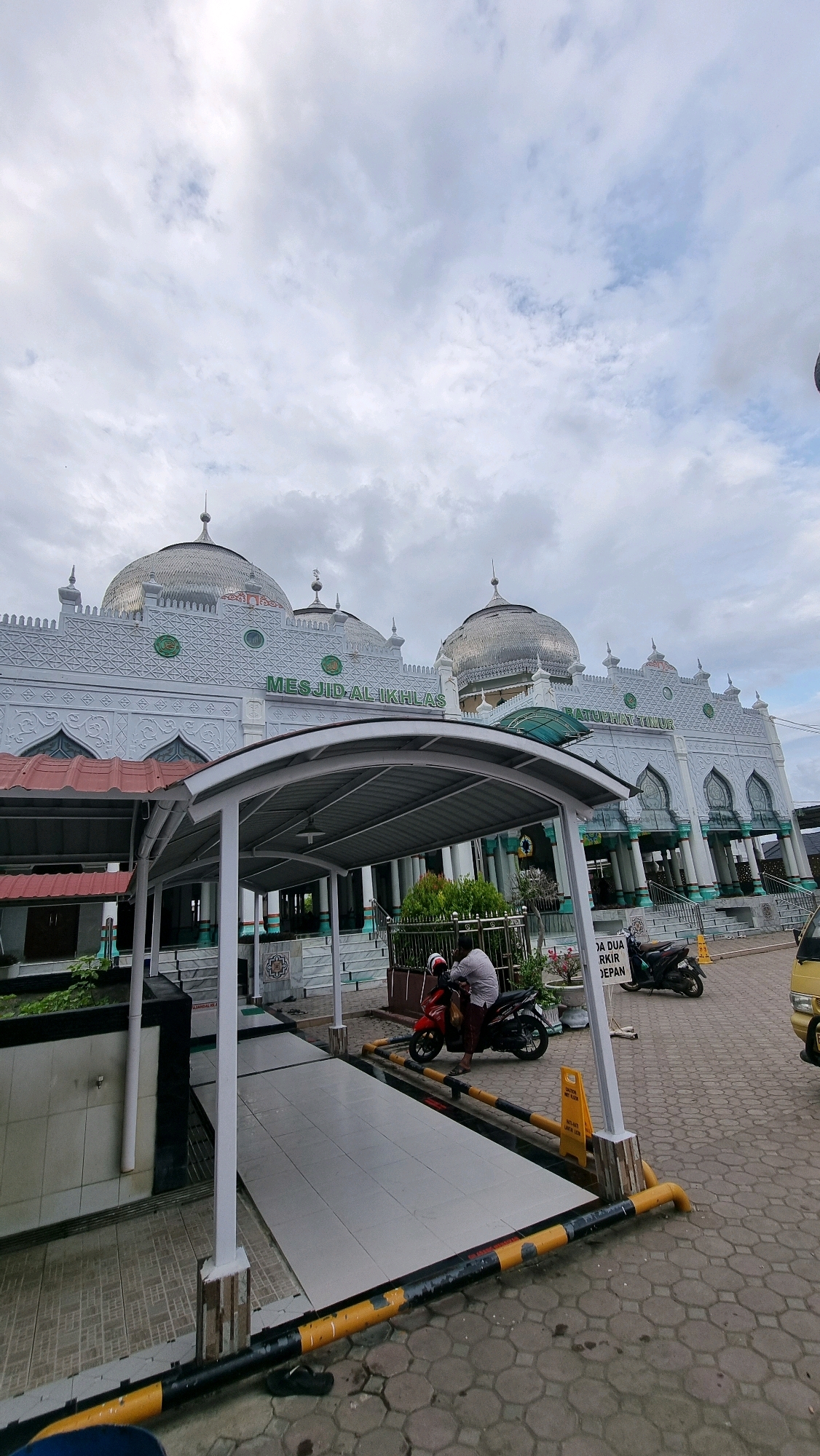 A wide shot captures the exterior of the Al-Ikhlas Mosque in Lhokseumawe, Indonesia, under a cloudy sky. The mosque is an ornate white structure with multiple domes, featuring intricate decorative patterns. A white covered walkway extends from the foreground towards the mosque entrance, with a ramp leading up to it. In the middle ground, a person sits on a red motorcycle parked in front of a low fence. To the right, another motorcycle is parked. A yellow cautionary sign stands near the motorcycles. The ground is paved with hexagonal stones. The image is taken from a low angle, emphasizing the mosque's architecture.