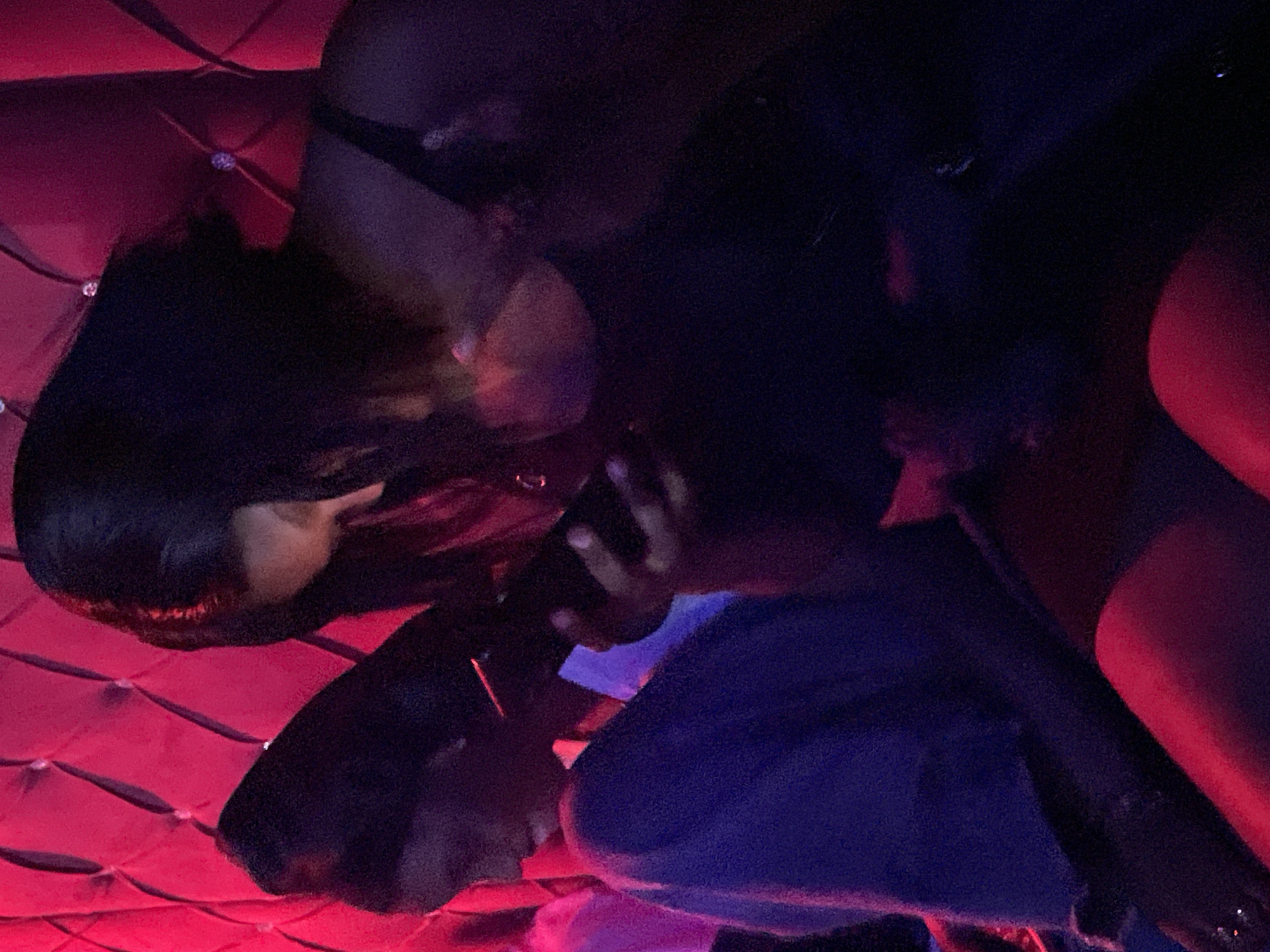 Two women are seated next to each other in a dimly lit room with plush red tufted upholstery. The woman on the left, wearing a dark blue garment, is looking down at her phone, which she holds in her hands. Her hair is dark and appears to be shoulder-length. The woman on the right, wearing a dark top, is also looking down at her phone. Her hair is dark and appears to be shorter, styled in a bob. Both women are seated close together, suggesting a shared activity or conversation, although their interaction with each other is not depicted. The overall lighting is red, with some purple and pink accents, possibly from ambient lighting in a nightclub or lounge. The exact location in the city of Durimi, Nigeria, cannot be determined from the image alone.