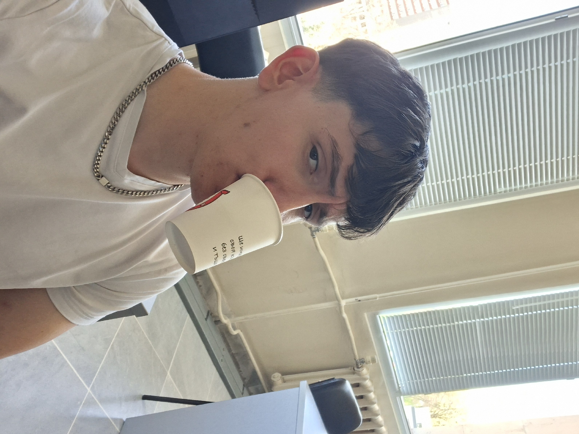 A close-up, low-angle shot shows a young man in a white t-shirt drinking from a white paper cup. He has dark hair, fair skin, and is wearing a silver chain. The cup obscures part of his face. Behind him, two windows with white blinds are visible. The lower window is open, and the outside is lit, hinting at daylight. The location is Unknown.