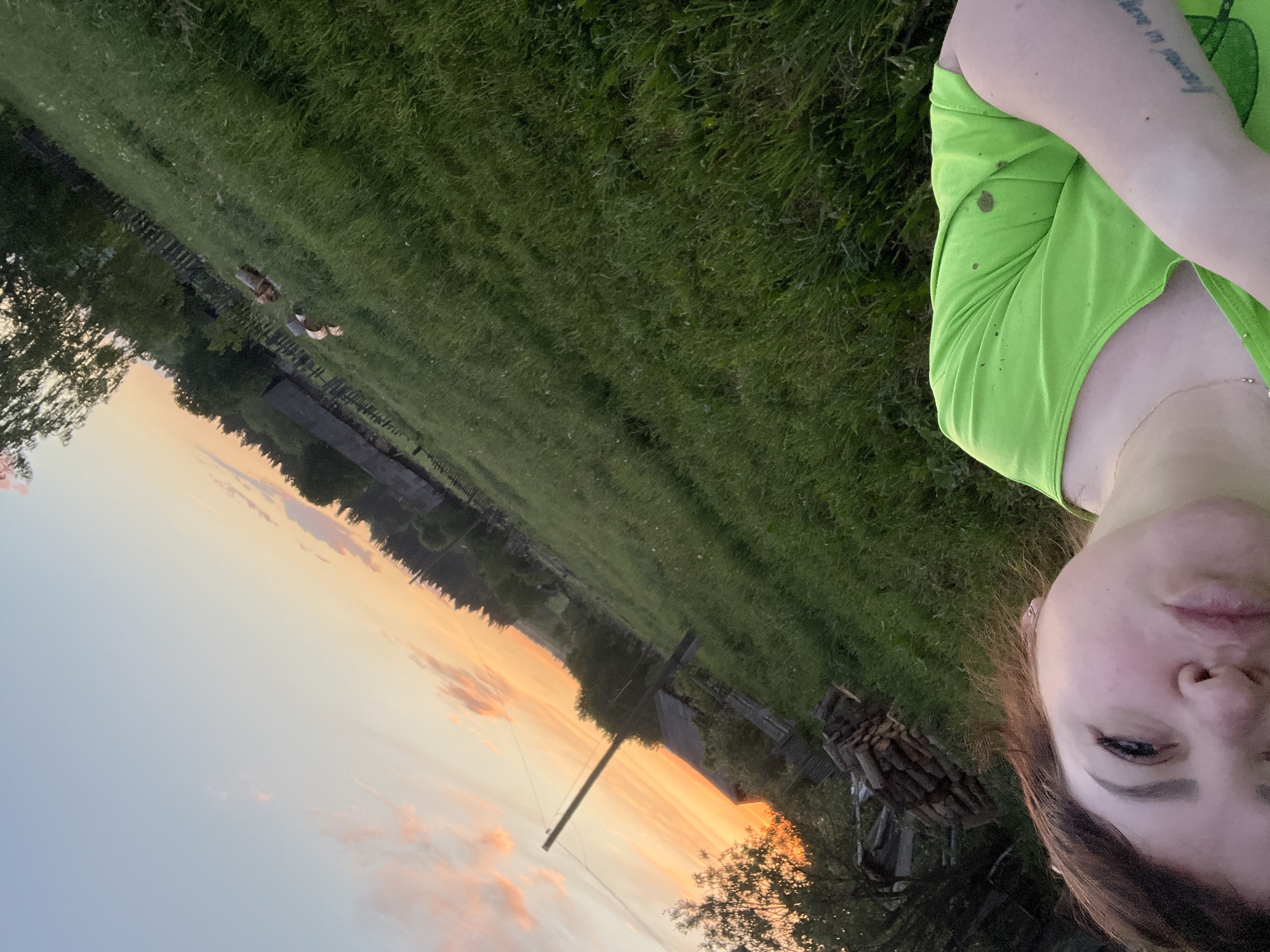 A daytime, selfie-style image depicts a fair-skinned female with brown hair, fair complexion, and wearing a bright green t-shirt. The subject's face fills the lower right quadrant of the frame. The subject is partially visible from the right side of the head, shoulder, and arm. On the upper part of the left arm is a tattooed phrase, partially obscured. A gold necklace is visible around the neck. The background shows a rural scene with a golden-orange sunset. A large open field of green grass occupies the upper portion of the image. In the mid-ground, trees, small buildings, and an old wooden fence line the horizon. A wooden utility pole is visible near the center of the frame. Piles of firewood are at the lower center-left. The image is rotated counterclockwise. The location might be Panteleevka, Russia, based on the request.