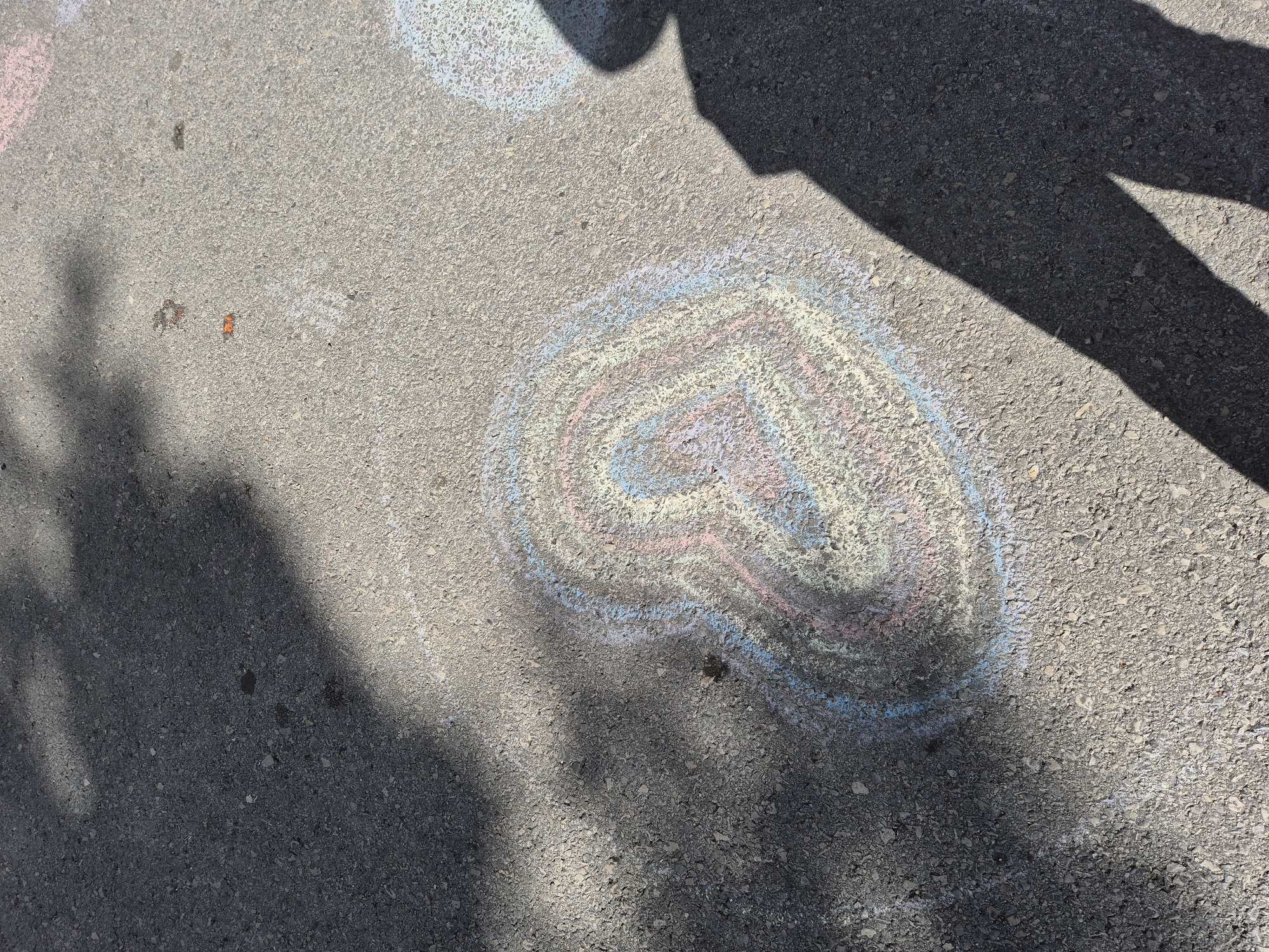 The image depicts a section of asphalt ground, likely a street or pavement. A multi-colored heart is drawn on the ground with chalk, exhibiting shades of blue, pink, and white. Shadows of a person and a tree branch are cast on the ground.