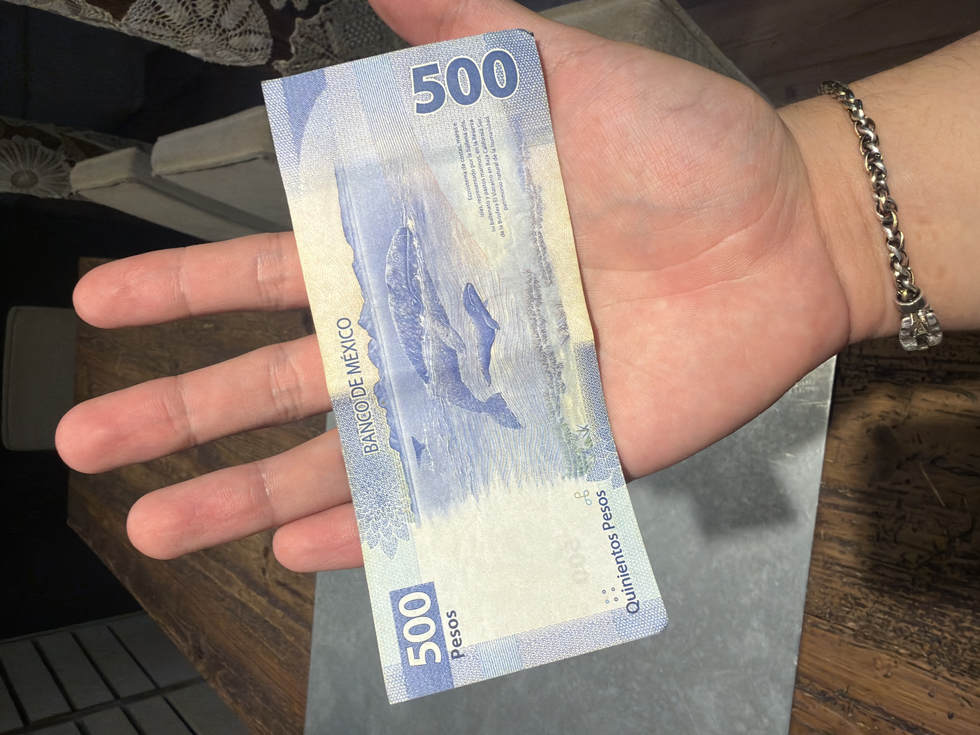 A person's hand holds a blue 500 Mexican pesos bill featuring whales and the inscription "Banco de Mexico". The bill also has the number 500 printed on it and the phrase "Quinientos Pesos" (Five Hundred Pesos). The person is wearing a silver chain-link bracelet on their wrist. The background includes a wooden surface, part of a chair, and a piece of lace fabric. The location is not Puebla, Mexico.