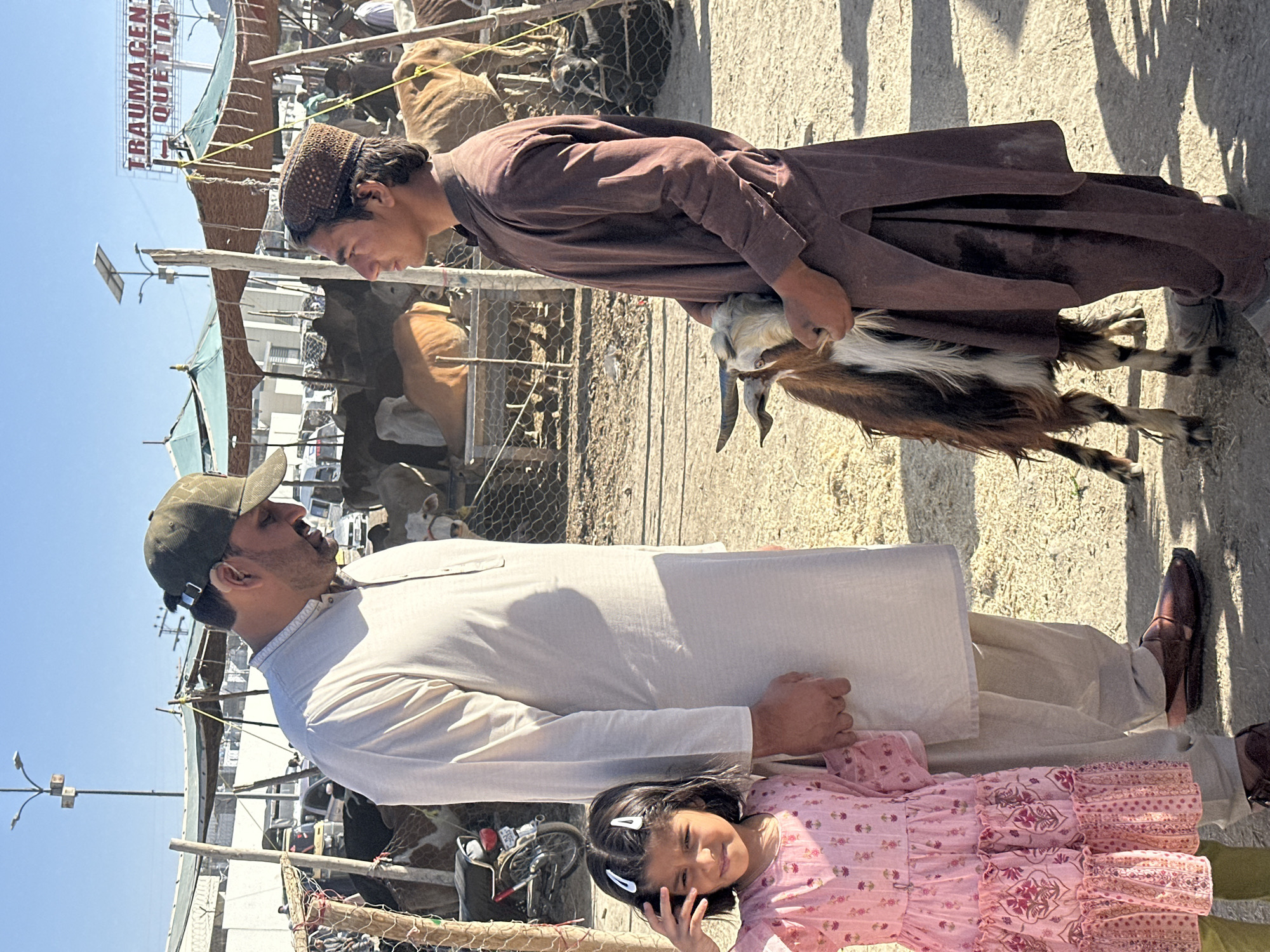 The image shows three people and a goat in an outdoor setting, likely a market or fair. In the background, there is a sign that reads "TRAUMA CENTRE QUETTA." The setting is crowded and suggests a bustling marketplace with animals and people.
A young man in a brown tunic is holding a goat, likely for sale or display. A man wearing a tan colored shirt and a baseball cap stands beside a small girl in a pink dress. The girl is making a gesture with her hand towards her face. The overall setting appears to be in Quetta City Tehsil, Pakistan, due to the sign visible.