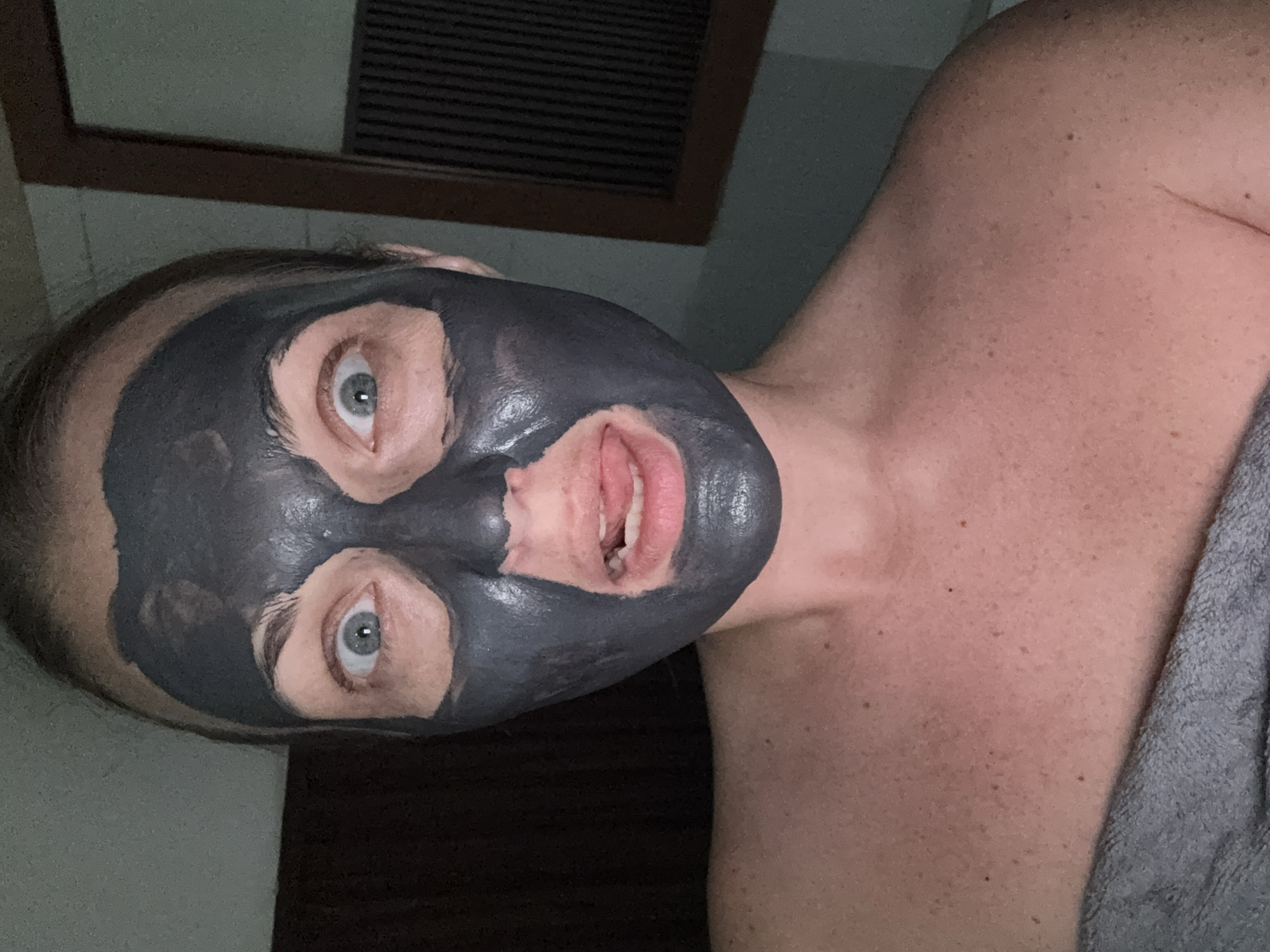 A woman with fair skin and light brown hair is applying a black charcoal face mask. She is looking at the camera with wide, surprised eyes and her mouth is open, with her tongue slightly sticking out. The mask covers most of her face, leaving her eyes and mouth visible. She is wearing a gray towel or wrap around her shoulders. The background is dimly lit and appears to be a bathroom, with a wooden cabinet or mirror on the right side of the image. The setting is an indoor location, and no specific landmarks of Denpasar, Indonesia are visible.