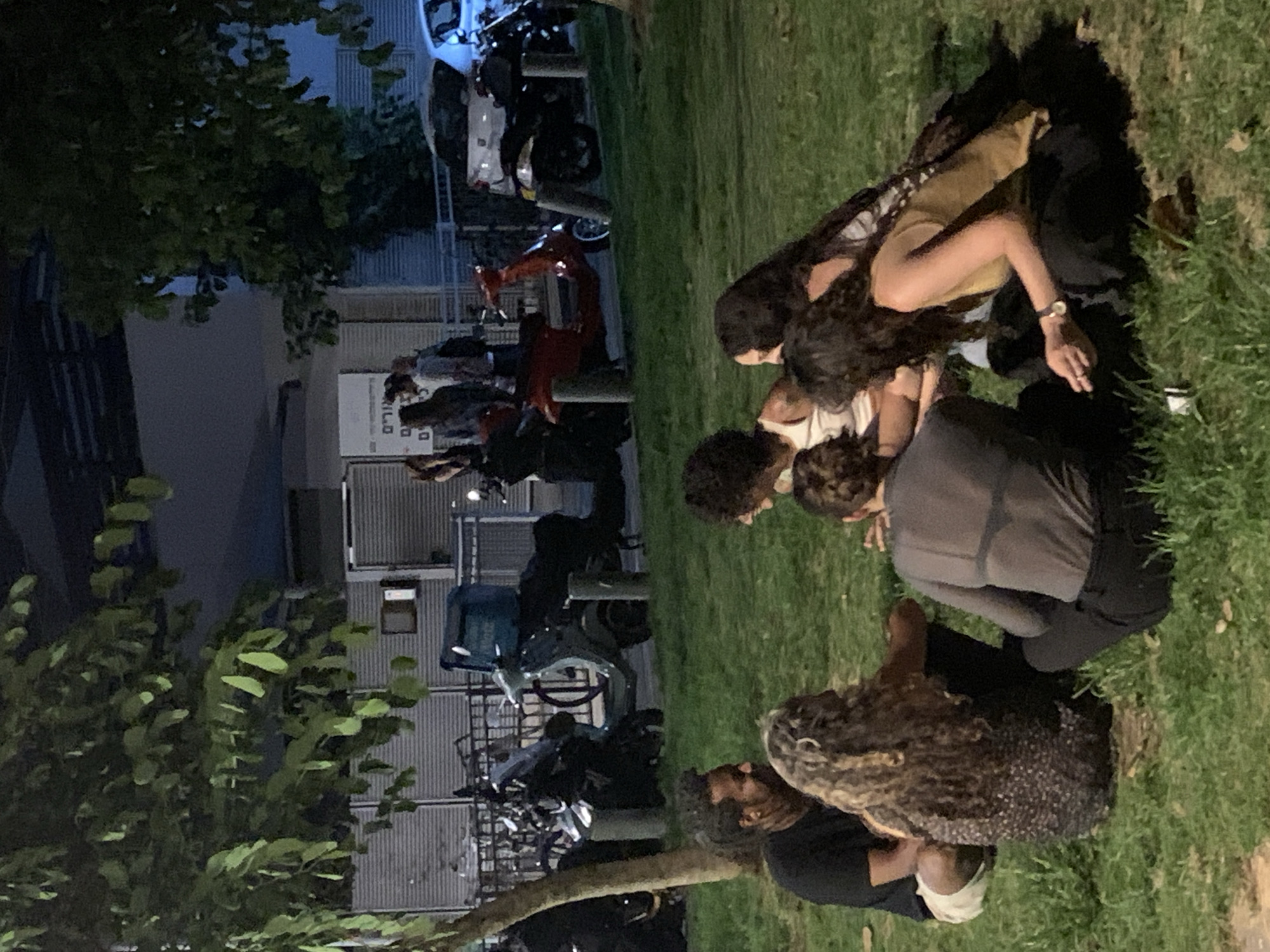 A group of six young adults are gathered on a grassy area in Tel Aviv at night. The group consists of four women and two men. They are sitting in a circle, facing each other, with their legs tucked underneath them.
The woman on the far left is wearing a black t-shirt and white shorts. She is looking to her right. Next to her, a woman with long, curly blonde hair is wearing a patterned dress. She is facing towards the center of the circle. In the center, a woman in a gray t-shirt with a black bra strap visible is facing away from the camera. To her right, a woman with long dark hair is wearing a yellow tank top and black pants. She is holding a cigarette in her right hand and looking down. Partially visible behind her is another woman with dark hair. The man on the far left of the circle is wearing a black t-shirt and light-colored shorts. He is looking at the woman with curly blonde hair. Another man, with short curly hair, is visible in the background, standing with a group of people.
In the background, several scooters are parked. One red scooter is in the foreground, and two other scooters, one dark and one lighter colored with a teal delivery box, are parked further back. A white car is also visible. The setting appears to be an urban area, possibly a park or public space, with trees providing shade and a building with shutters in the background. The lighting suggests it is nighttime, with some artificial lights illuminating the scene.