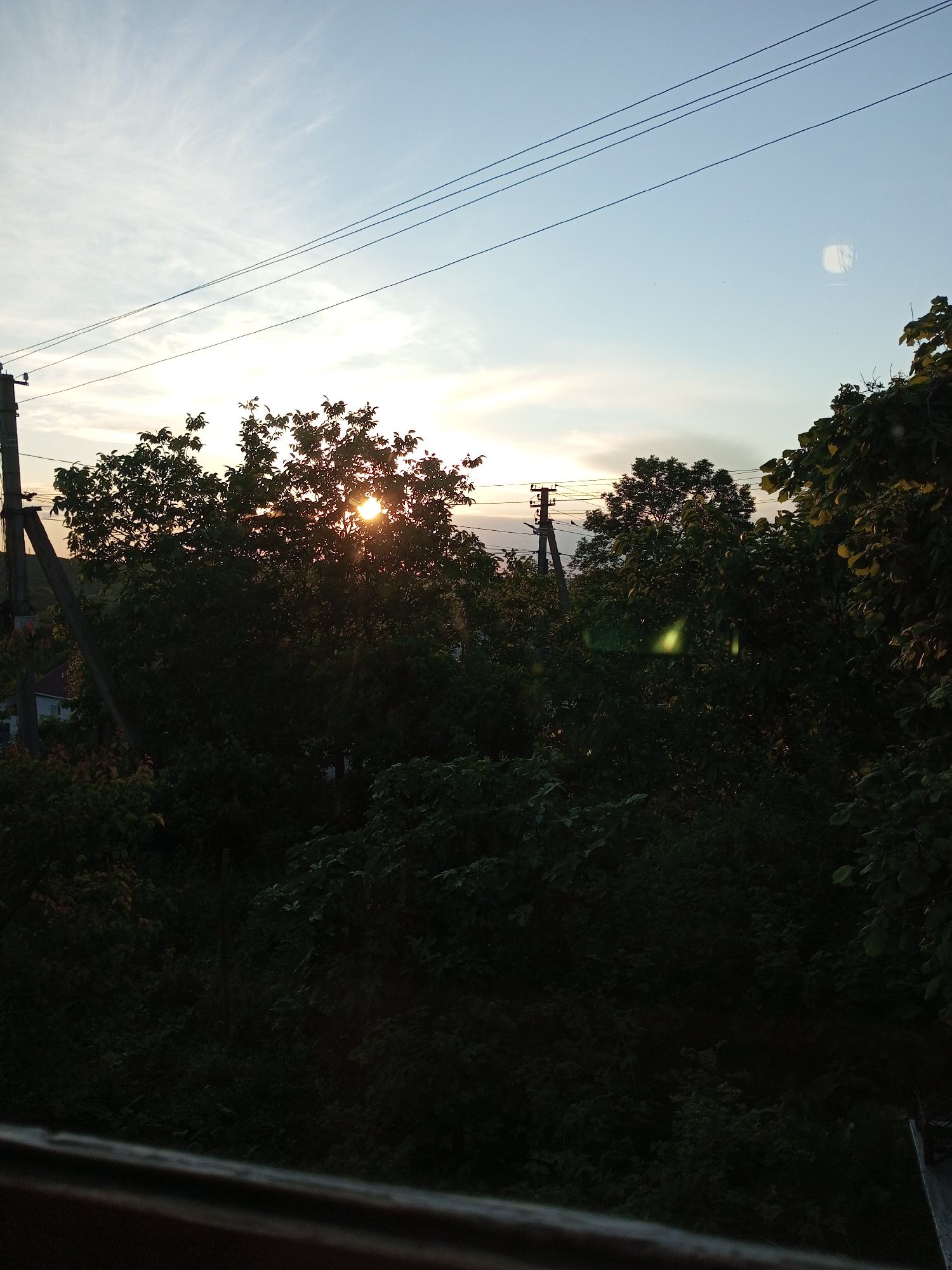 The image depicts a view of a tree line against a sky during sunset. The sun is partially obscured by a large tree in the center, casting rays of light through its leaves. The sky is a gradient of pale blue to a lighter hue near the horizon, with wispy clouds. Power lines are visible stretching across the upper portion of the frame. In the foreground, there is a wooden railing or surface. The scene appears to be outdoors, possibly in a rural or suburban setting, given the presence of trees and power lines. There is a reflection or lens flare in the upper right portion of the sky.