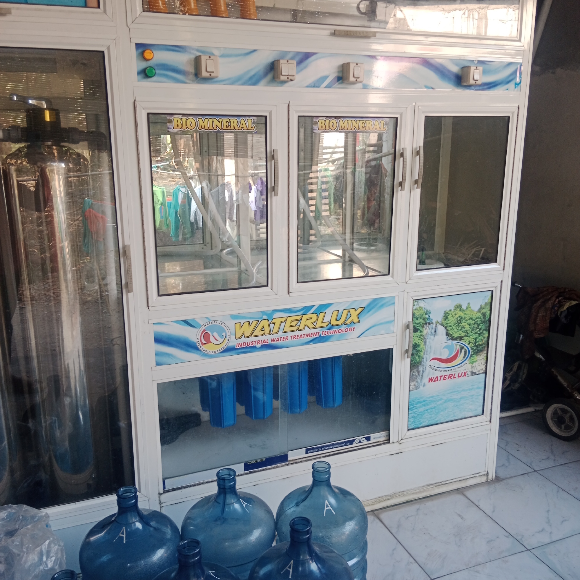 This is a medium-shot, eye-level, full-body photograph of a water dispenser machine in Tegowanu Kulon, Indonesia. The machine is white with glass doors and features various labels and buttons. On the top portion of the machine, there are three square light switches and two circular indicator lights, one orange and one green. Below the switches, a blue and white wave design is visible. Two upper glass doors display the text "BIO MINERAL" in yellow lettering. A lower section of the machine has a larger glass panel with a blue and white banner that reads "WATERLUX INDUSTRIAL WATER TREATMENT TECHNOLOGY" and a smaller glass panel with a picture of a waterfall and the "WATERLUX" logo. The bottom section of the machine contains blue water filter cartridges visible through glass. In the foreground, several large blue plastic water jugs are arranged, some with the letter "A" written on them in white. To the left of the machine, a large stainless steel water filtration tank is visible. To the right of the machine, a dark-colored stroller or cart with a white wheel is partially visible. The floor is tiled with a light grey and white pattern. The background is blurred but appears to be an outdoor or semi-outdoor setting with some vegetation and laundry hanging. The overall scene suggests a commercial or residential area where water dispensing services are offered.