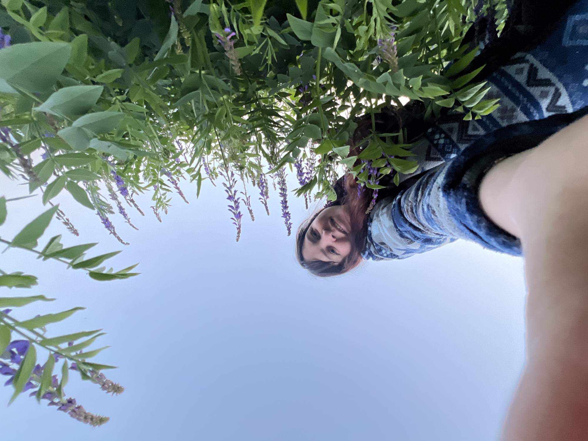 A selfie captured from a low angle, showing a woman with long dark hair and a blue patterned sweater looking up at the camera with a smile. She is in a field of green plants with small purple flowers. The sky is clear and pale blue. The photo appears to be taken in Panteleevka, Russia.