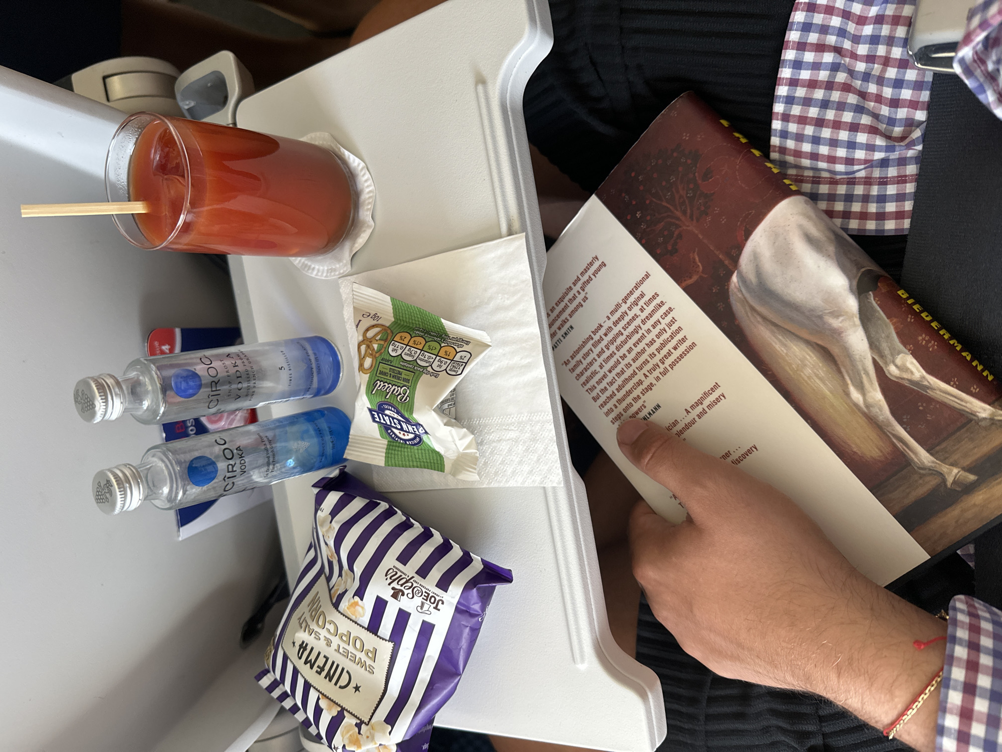 An overhead medium shot shows a person's arm and hand resting on a book on a tray table aboard an airplane. The tray table holds two small bottles of Ciroc Vodka, a bag of Cinema Sweet & Salty Popcorn, a bag of Penn State Baked Pretzels, and a glass of tomato juice with ice and a wooden stirrer. The book is open, and the person's right hand is holding it. The person is wearing a plaid shirt with red and blue checks. The background is blurred but appears to be the interior of an airplane cabin. The overall scene suggests a traveler enjoying a drink and a snack while reading on a flight.