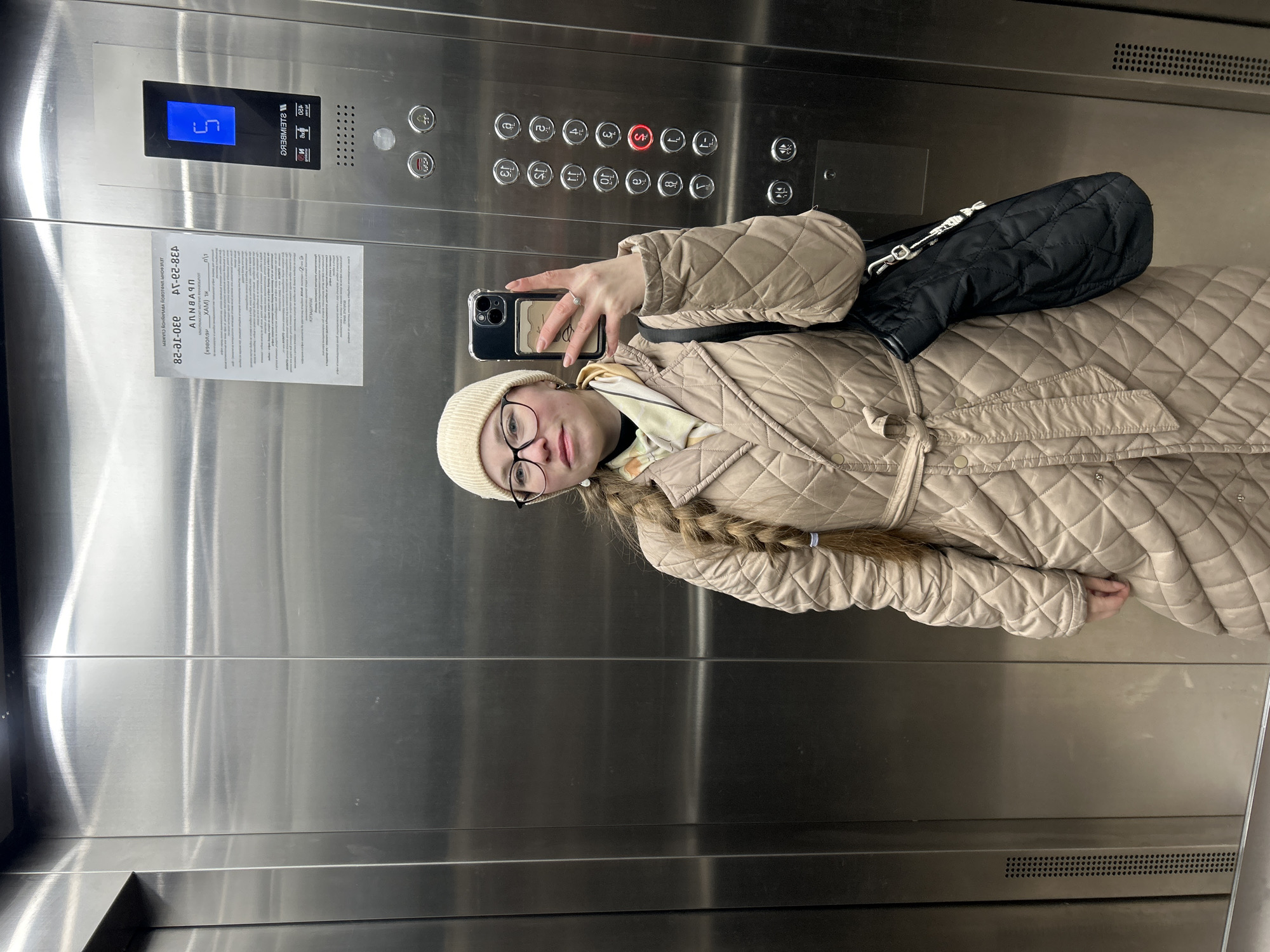 A young woman with long blonde braided hair, wearing glasses, a beige beanie, and a quilted beige jacket, takes a mirror selfie inside an elevator. She holds a smartphone with a black case in her right hand, revealing a ring on her finger. A black quilted shoulder bag is visible over her left shoulder.
The elevator interior features brushed stainless steel walls, reflecting the woman and the cabin's lighting. The digital display above the control panel shows the number "5" in blue, indicating the current floor. The control panel has numerous silver buttons for floors 1 through 16, with the "S" button illuminated red. Other buttons for elevator functions (door open/close, alarm) are also visible.
A printed notice in Russian is taped to the elevator wall. The text, in mirror image, informs residents of a hot water shutdown due to electrical equipment maintenance and replacement of water risers, scheduled from March 20, 2023, to March 28, 2023. It includes emergency service contact numbers and the name of the management company, "ООО "УК "УПРАВДОМ"". This, along with the Russian text, places the location contextually in Saint Petersburg, Russia. The woman's attire suggests cooler weather, possibly late winter or early spring. She maintains a neutral expression, looking directly at the phone.